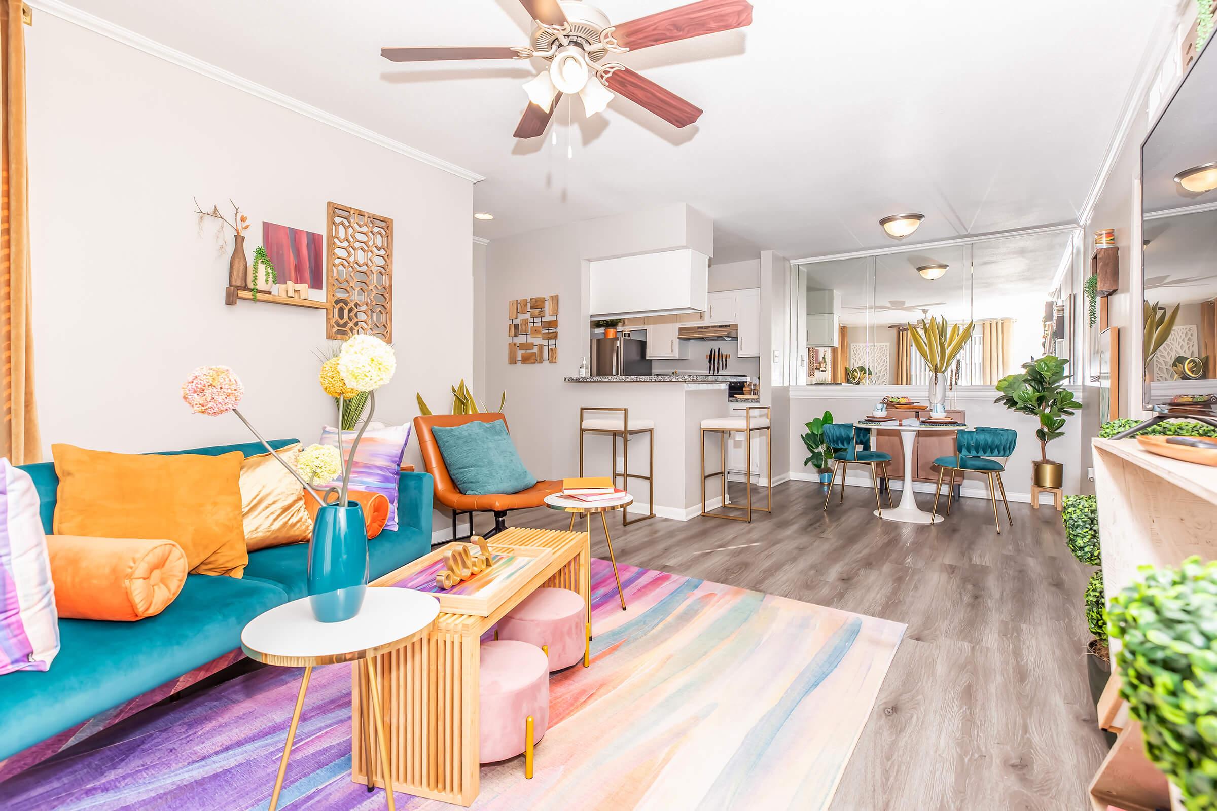 A cozy living room featuring a colorful sofa with decorative cushions, a modern coffee table, and a vibrant area rug. There are stylish chairs, a dining area, and plants throughout the space. Large windows let in natural light, creating an inviting atmosphere.