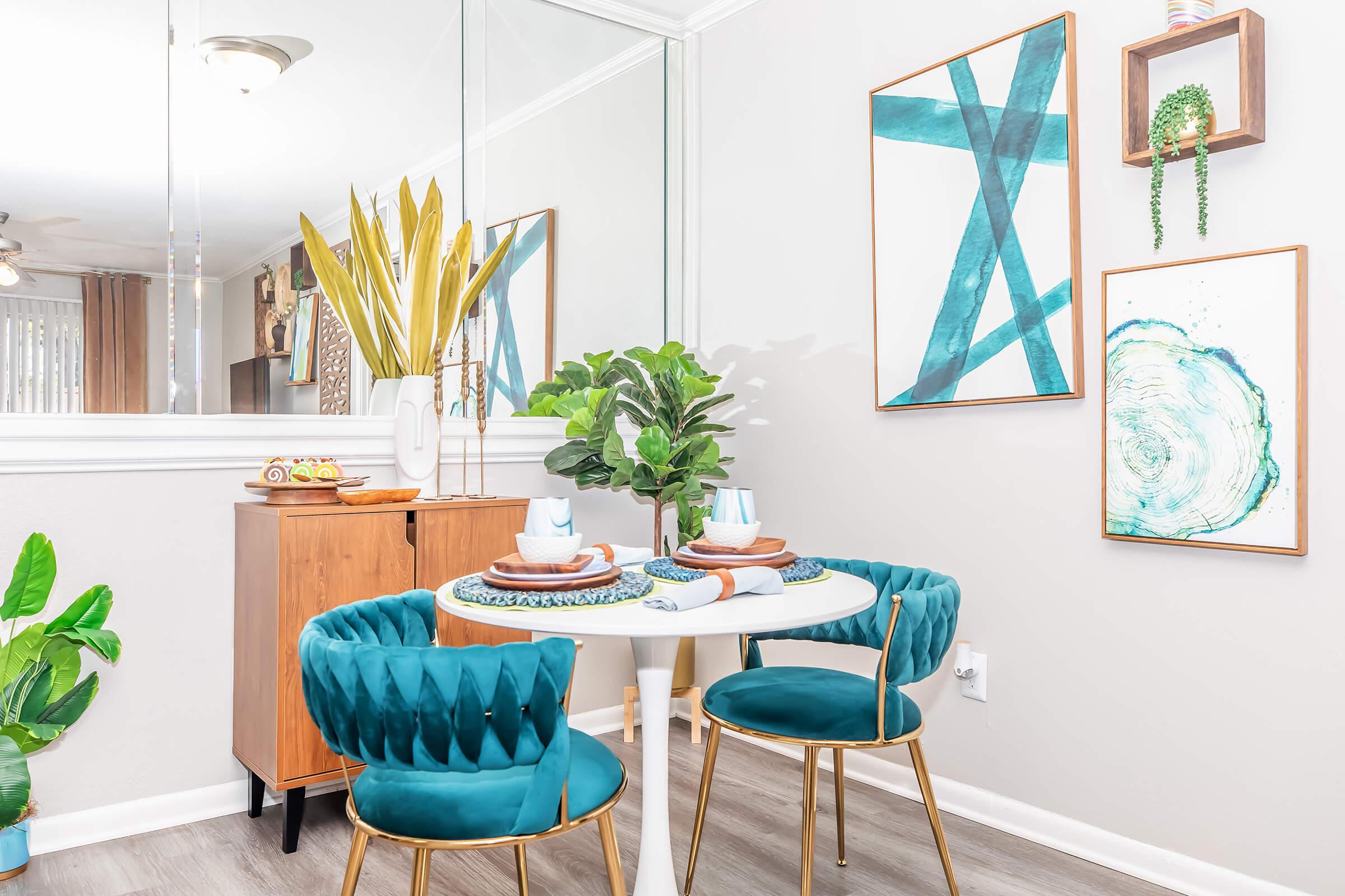A stylish dining area featuring a round white table with turquoise upholstered chairs. The table is set with plates and utensils, surrounded by greenery. A wooden cabinet and decorative wall art are visible, including abstract designs and a plant in a vase. A large mirror reflects the space, enhancing its brightness.