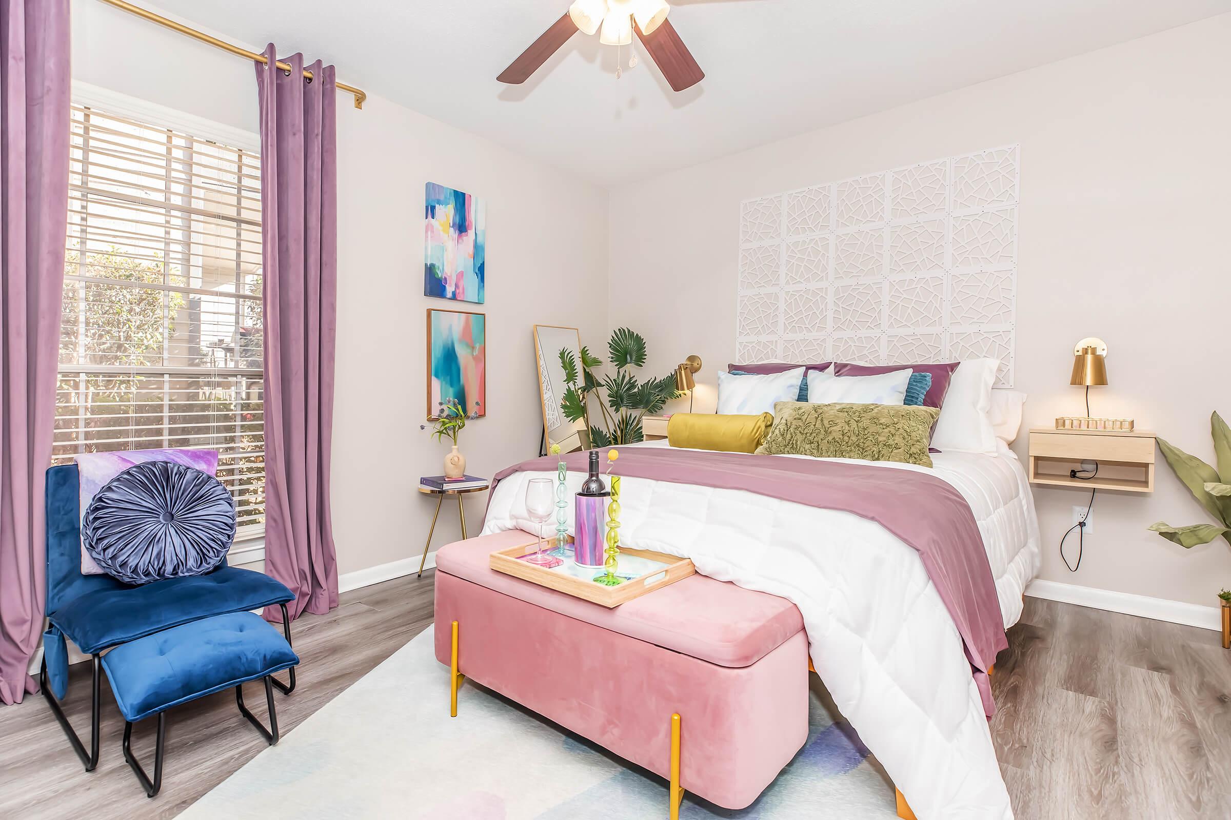 A cozy bedroom featuring a queen-sized bed with decorative pillows and a textured headboard. A pink ottoman sits at the foot of the bed, accompanied by a small side table and a lamp. The room is adorned with plants, colorful artwork on the walls, and layered curtains framing a window that allows natural light to enter.