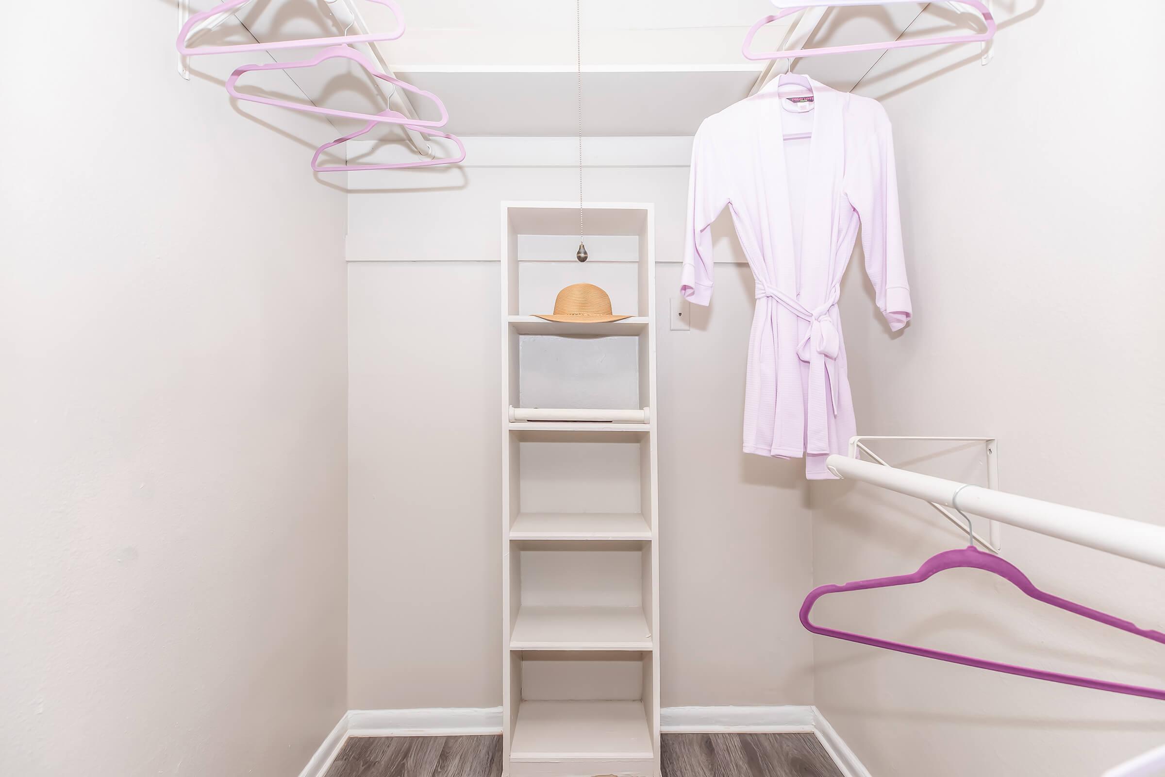 A minimalist closet with light gray walls, featuring a white shelving unit, light pink hangers, and a lavender robe hanging on the side. A brown hat is placed on one of the shelves, adding a touch of warmth to the space. The floor is a wood-like material, creating a clean and organized appearance.