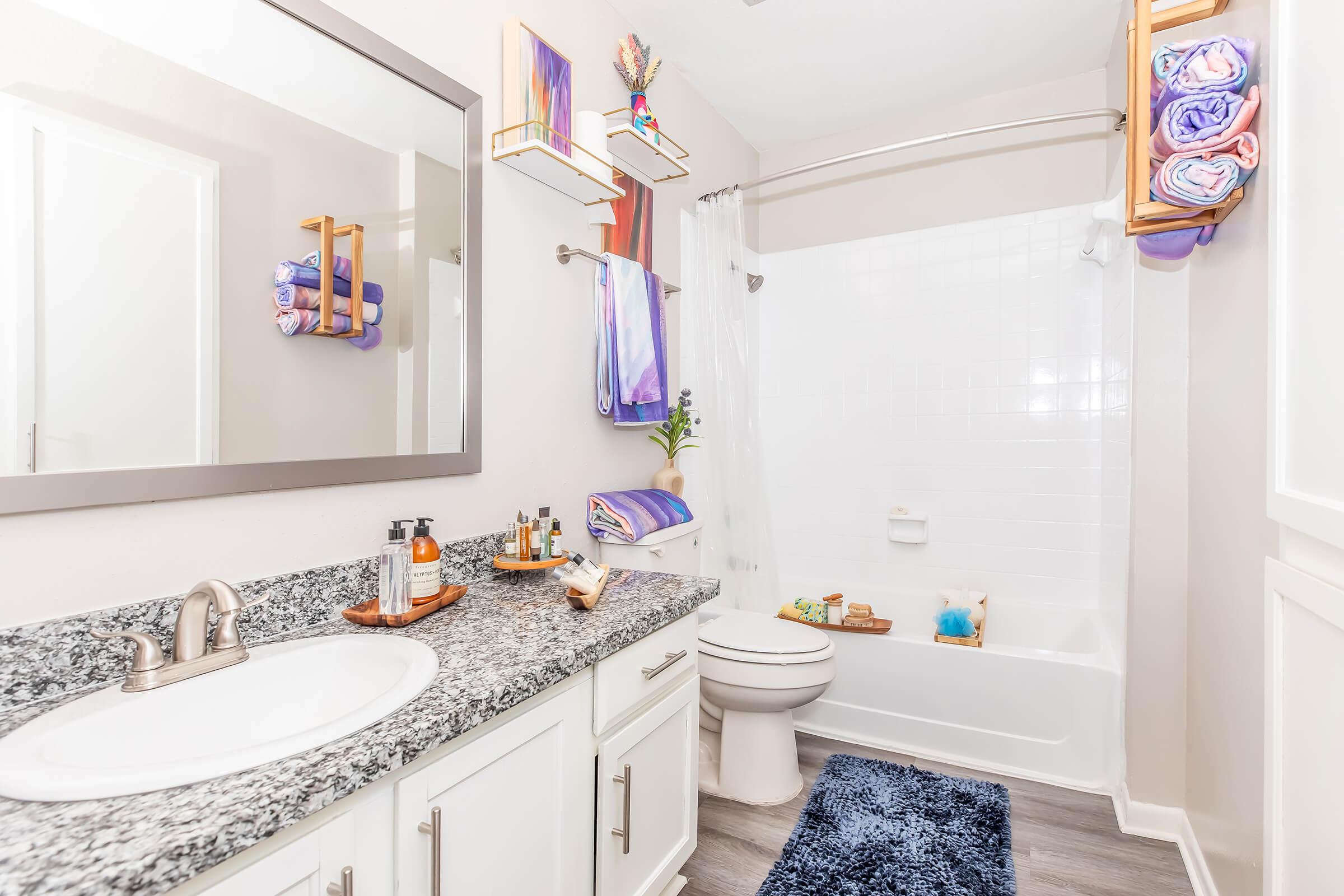 Bright and clean bathroom featuring a white bathtub with a shower, a granite countertop with toiletries, and neatly arranged towels on a wall-mounted rack. A large mirror reflects the well-lit space, enhancing the modern aesthetic with shades of purple and neutral tones. A plush rug adds a cozy touch.