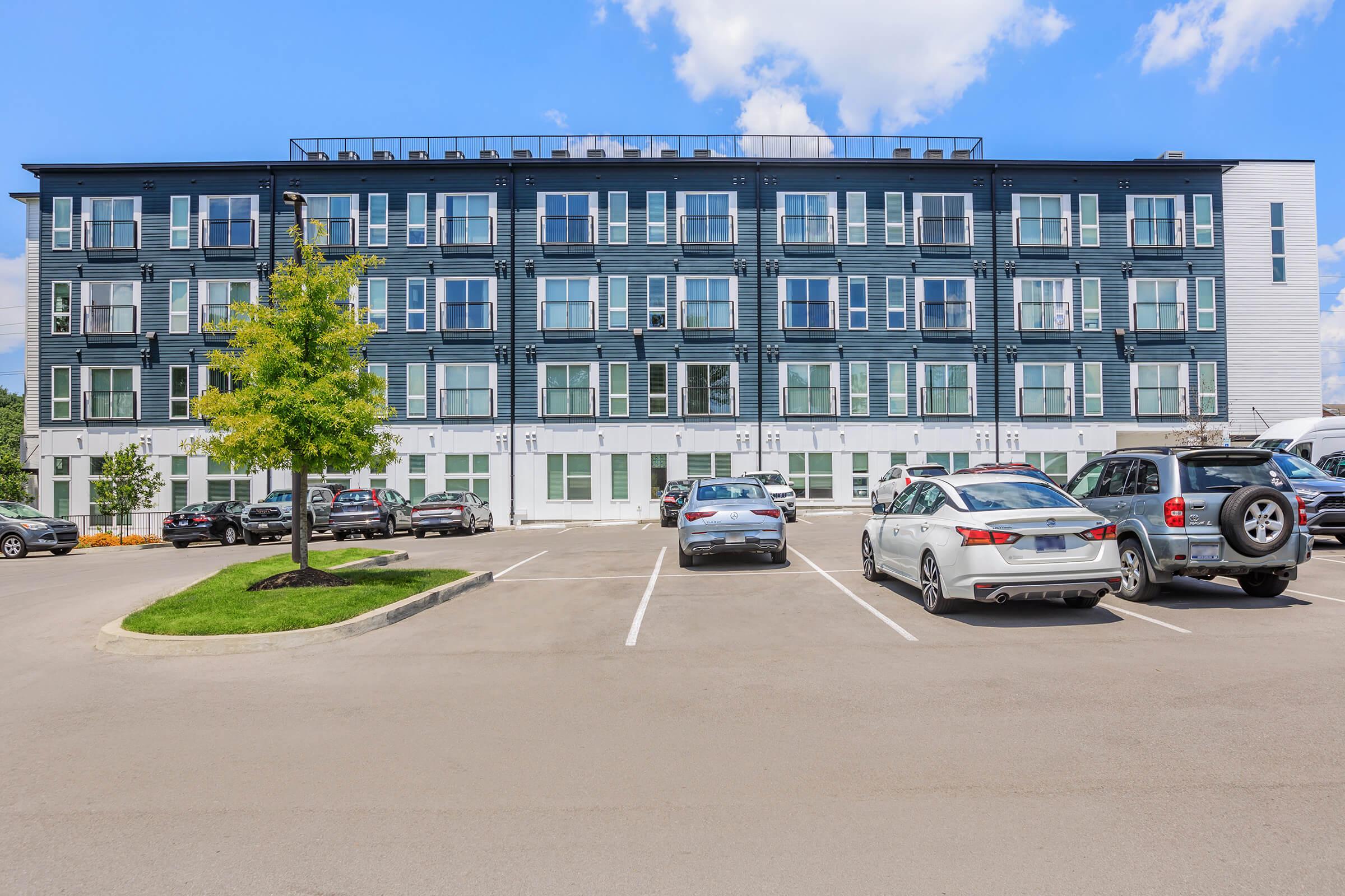 Modern multi-story building with a sleek design featuring large windows and balconies. Surrounding parking lot with several parked cars. Bright blue sky with a few clouds visible above.