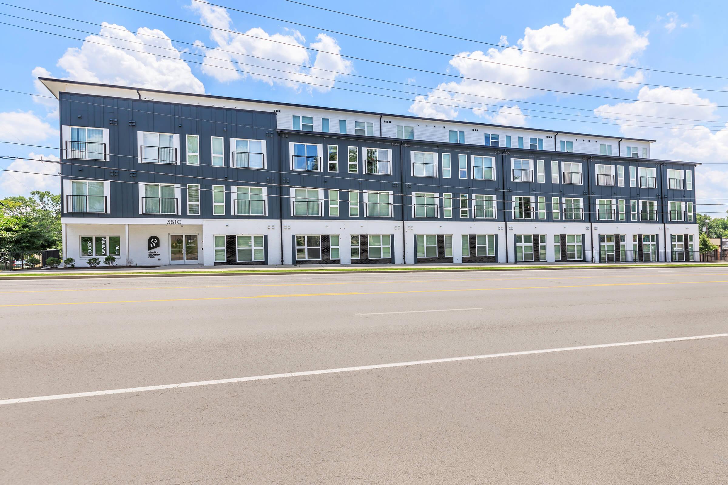 Modern multi-story building featuring a sleek, contemporary design with large windows. The façade showcases a mix of grey and white colors, and the structure spans several floors. A blue sky with fluffy clouds is visible in the background, along with a road in the foreground.