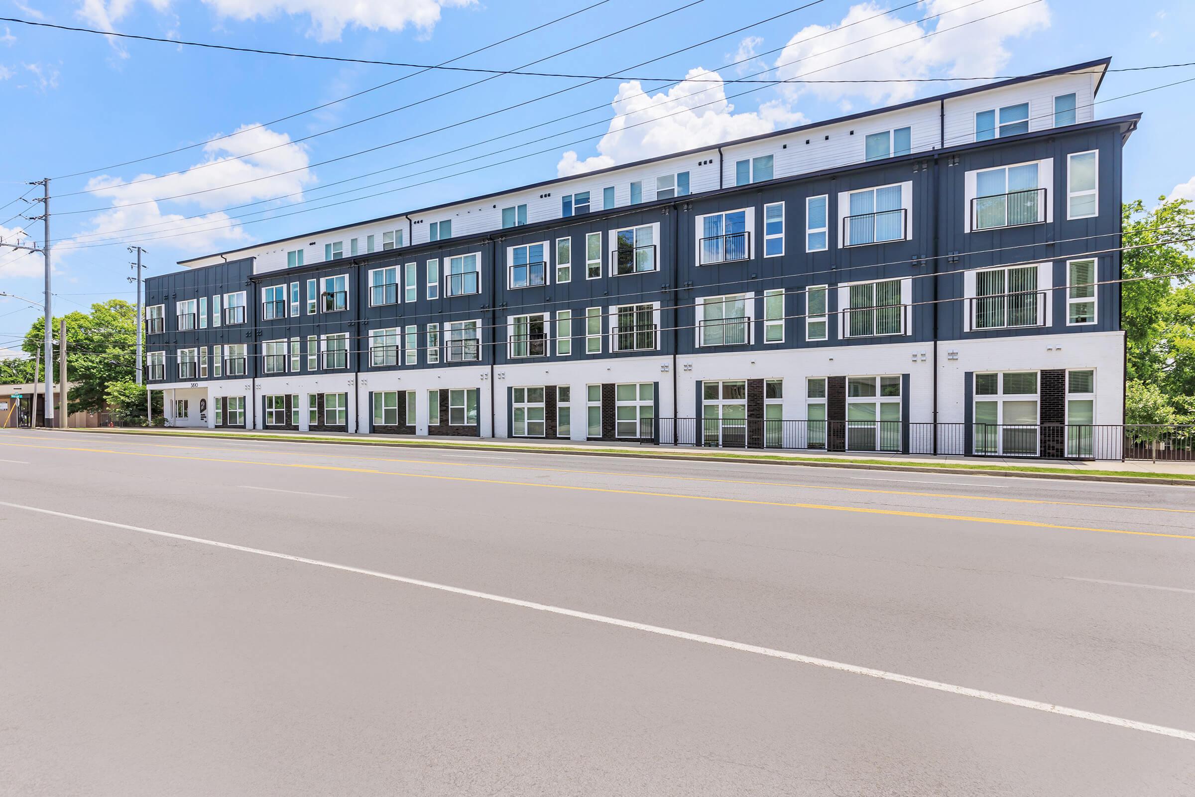Modern multi-story apartment building with large windows and a sleek black and white exterior, situated along a wide road. Clear blue sky with a few clouds overhead, and utility poles along the street. The building features balconies on the upper floors.