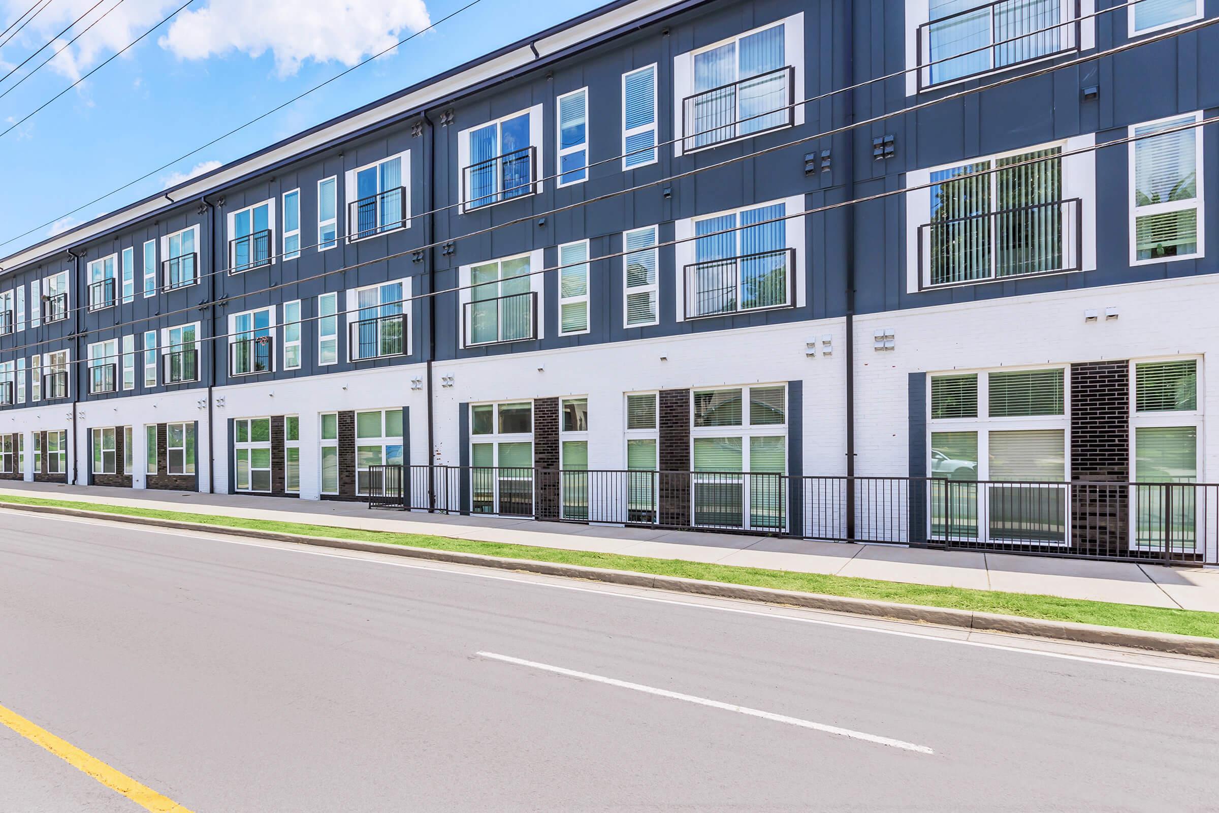 A modern, two-story building with a sleek black and white exterior. It features multiple large windows, balconies, and a clean sidewalk alongside the street. The scene is set on a clear day with blue skies and sparse clouds, showcasing a contemporary urban residential design.
