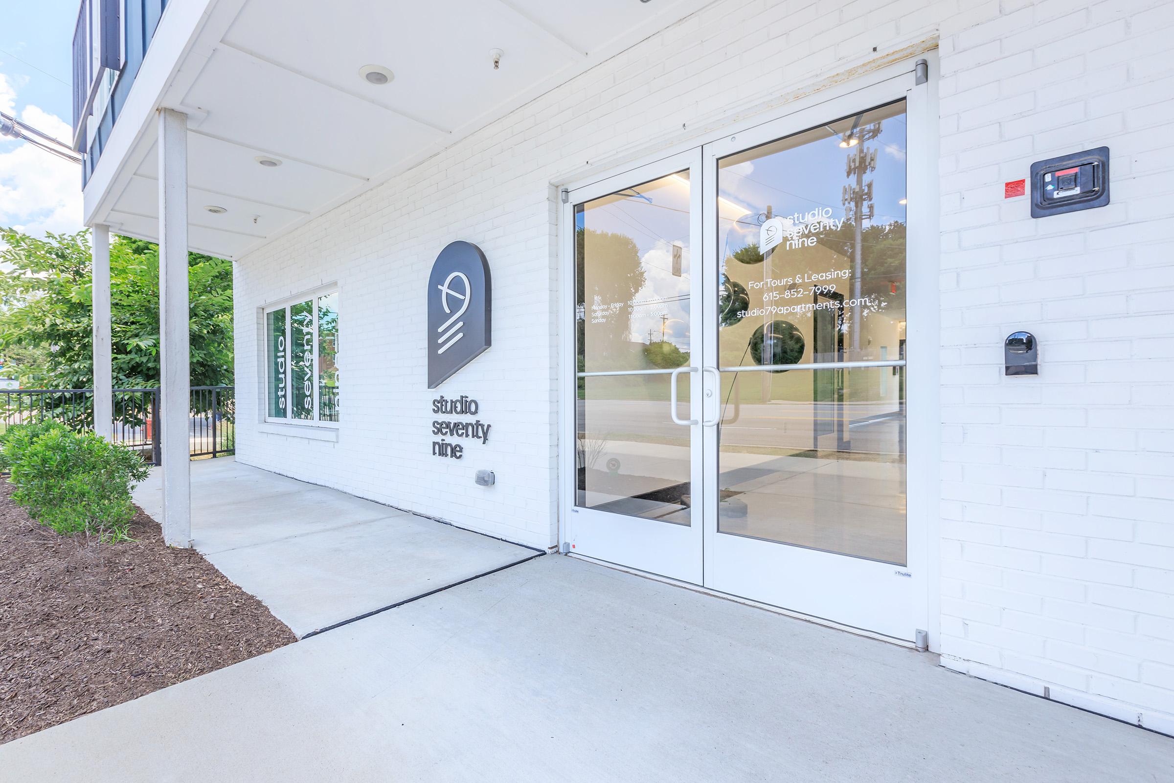 Entrance to Studio Seventy Nine featuring large glass doors and a modern logo on the brick wall. The pathway is paved, with greenery on one side and a small plant bed. The building has a contemporary design, with large windows allowing natural light inside.