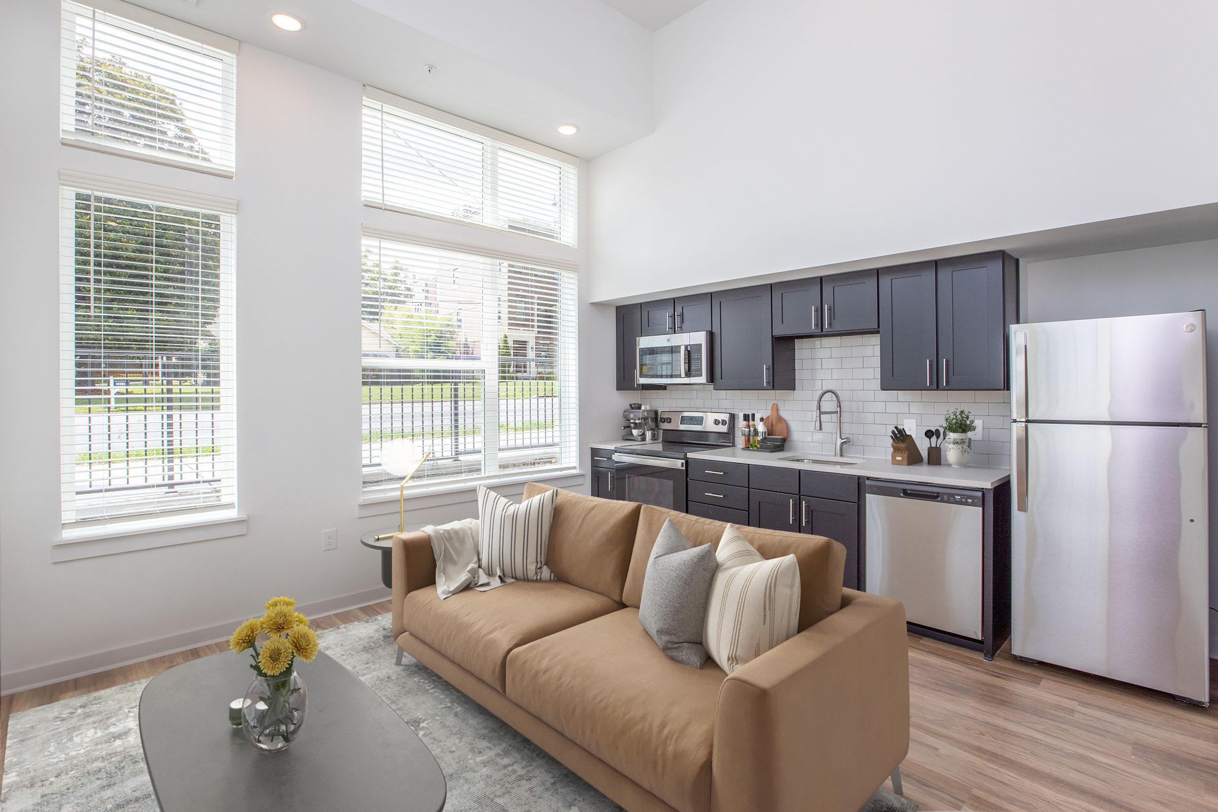 A modern living room featuring a brown couch with decorative pillows, a coffee table with yellow flowers, and an open kitchen with dark cabinets. Large windows allow natural light to fill the space, showcasing stainless steel appliances and a sleek design. The overall ambiance is bright and inviting.