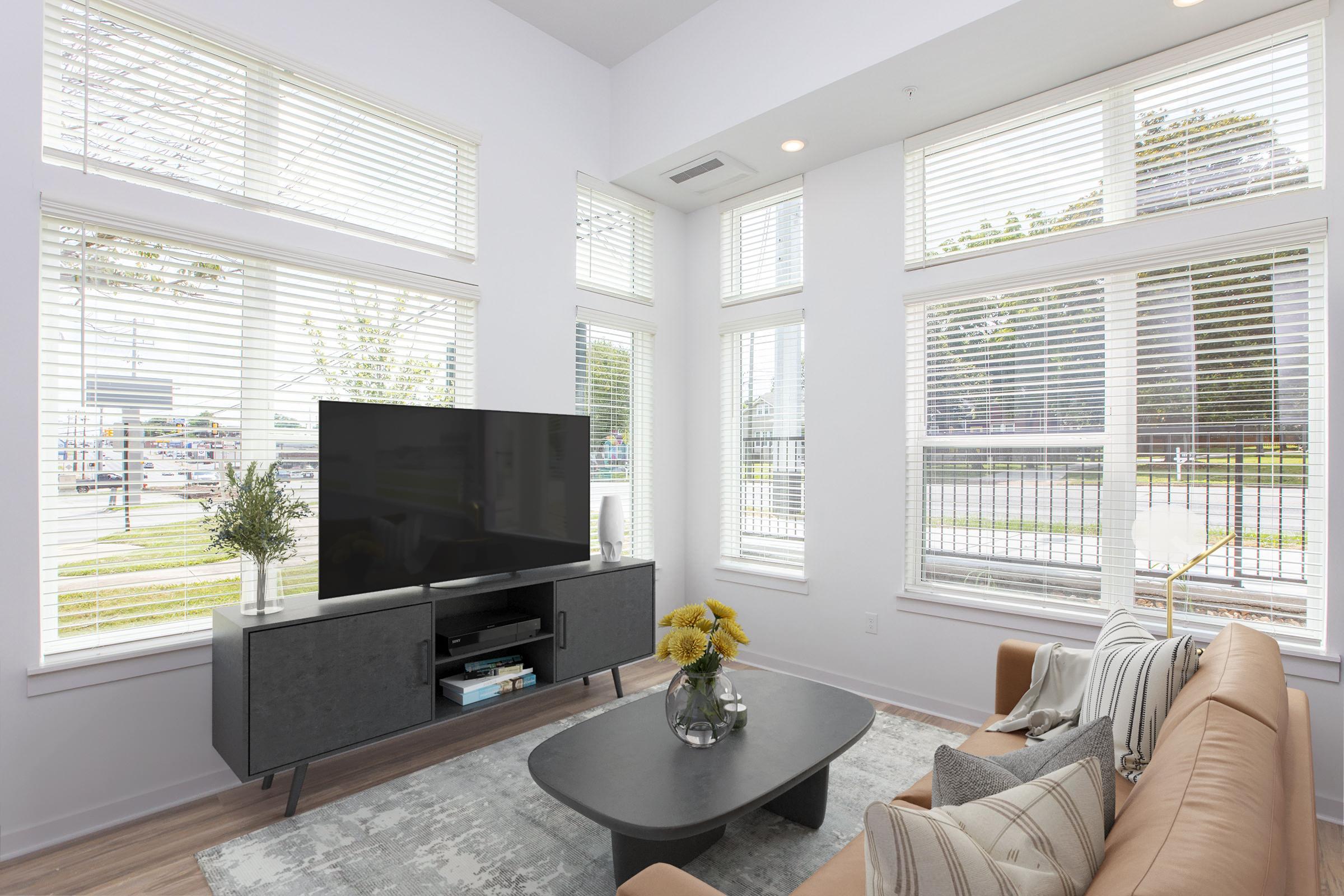 Modern living room featuring large windows with blinds, a flat-screen TV on a dark wooden console, a round coffee table with a vase of yellow flowers, and a tan sofa adorned with decorative pillows. The room has a light color palette and a cozy, inviting atmosphere.
