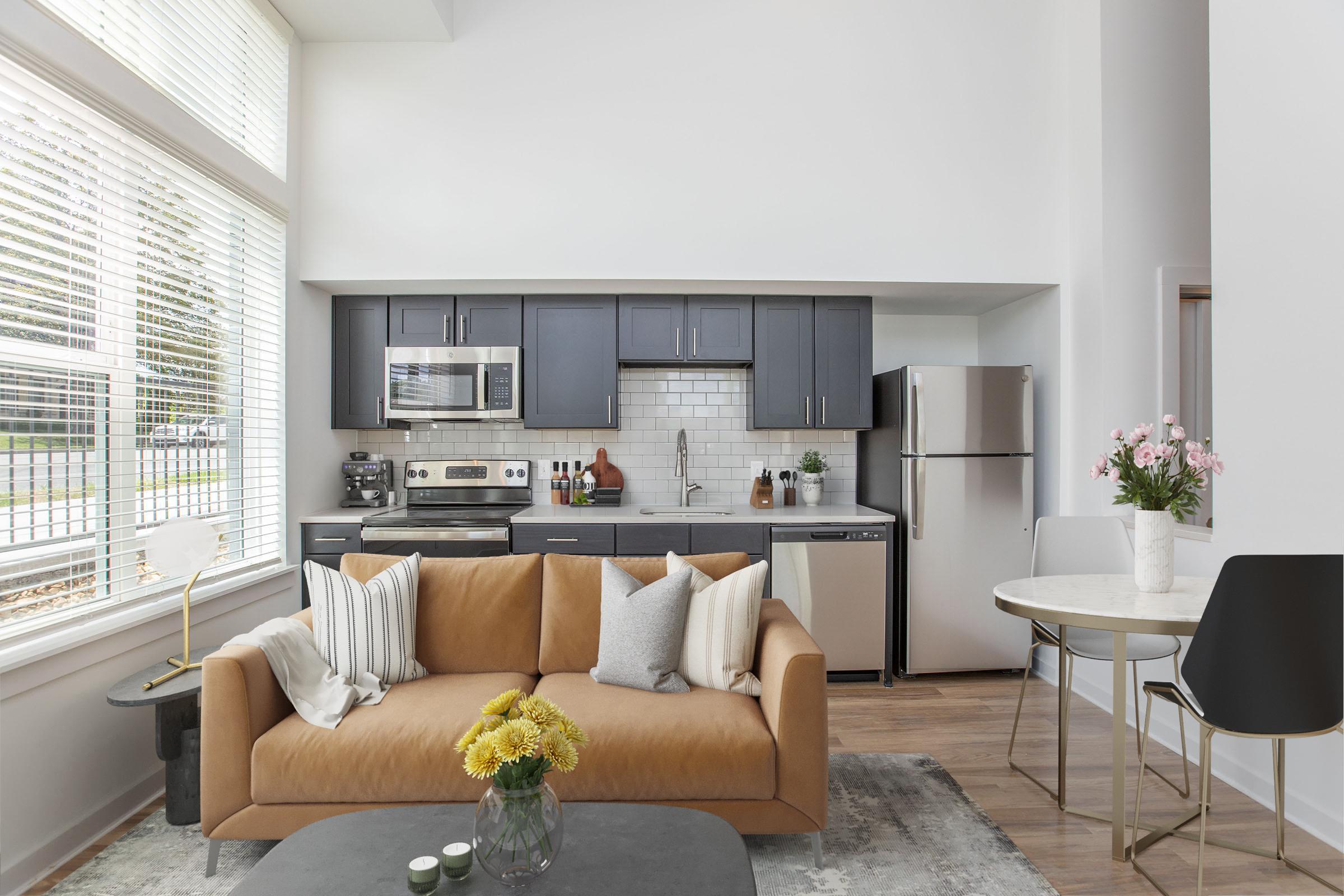 A modern living space featuring a cozy brown sofa with decorative pillows, a round coffee table, and a dining area with a small table and chairs. The kitchen in the background has dark cabinets, stainless steel appliances, and a white tile backsplash, with large windows allowing natural light to enter. Fresh flowers are placed on the table.