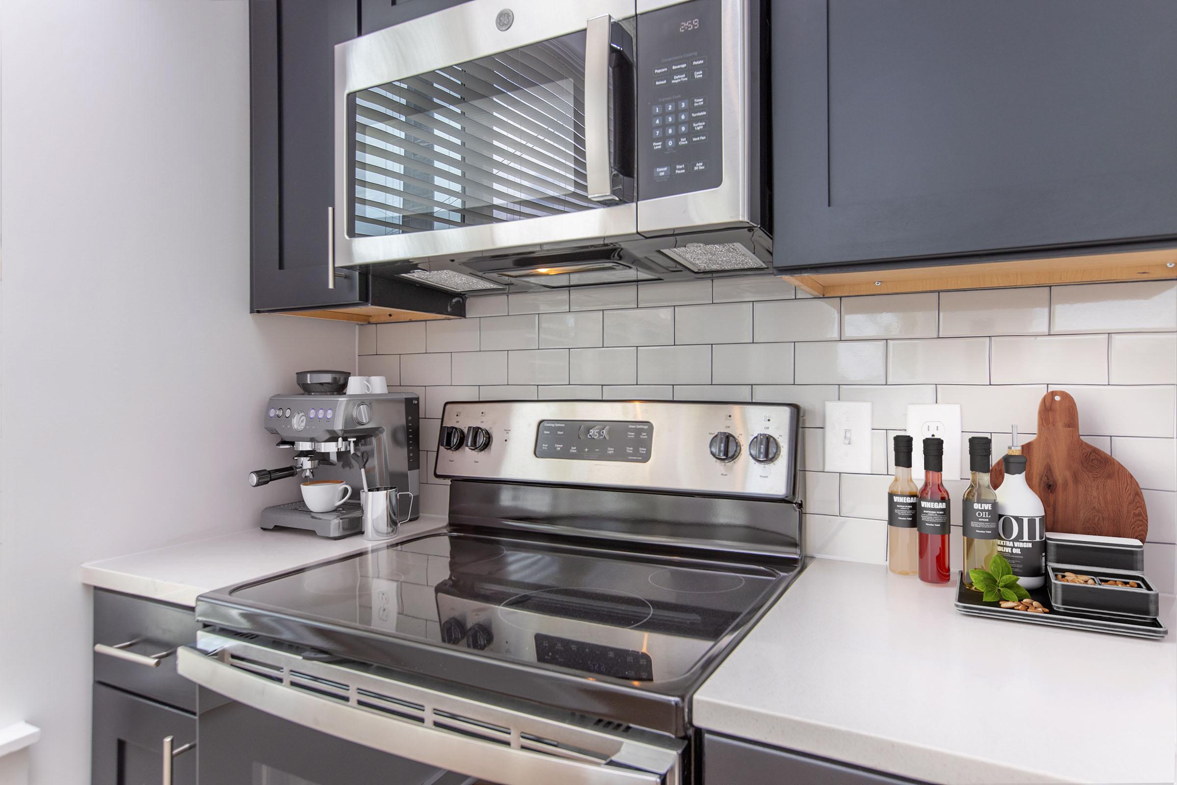 A modern kitchen featuring a stainless steel microwave above a stove, with a sleek countertop and dark cabinetry. An espresso machine and accessories are on the side, along with a wooden cutting board and small plants for decoration. The backsplash is white subway tile, complementing the contemporary design.