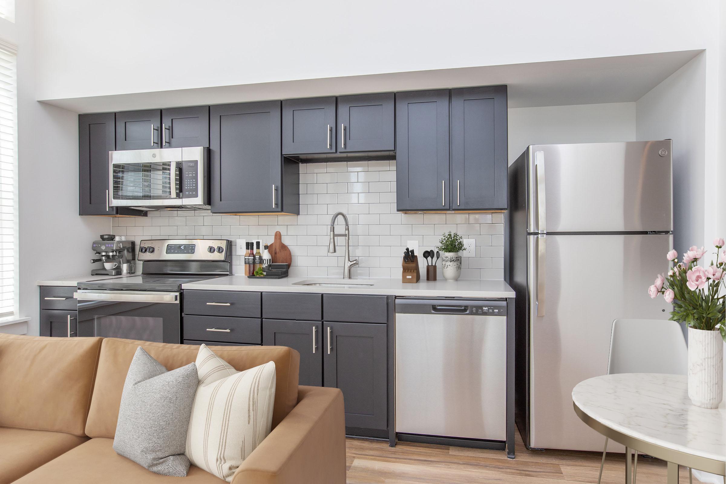 A modern kitchen featuring dark cabinets, a stainless steel refrigerator, a microwave, and a sleek sink. The countertop has decorative items and a potted plant. In the foreground, there’s a comfortable beige couch with decorative pillows and a round table with a vase of pink flowers. Bright natural light fills the space.