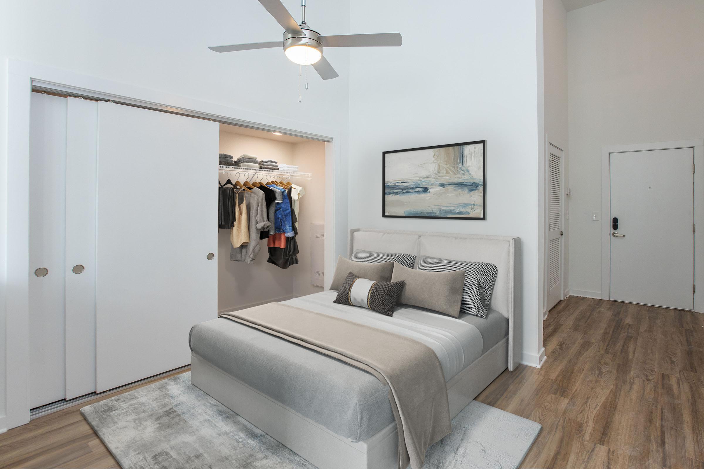 A modern bedroom featuring a neatly made bed with gray and white bedding, a light gray upholstered headboard, and decorative pillows. A ceiling fan is visible above, while a closet with sliding doors displays neatly arranged clothing. A piece of abstract wall art hangs above the bed, and a doorway is visible leading outside.