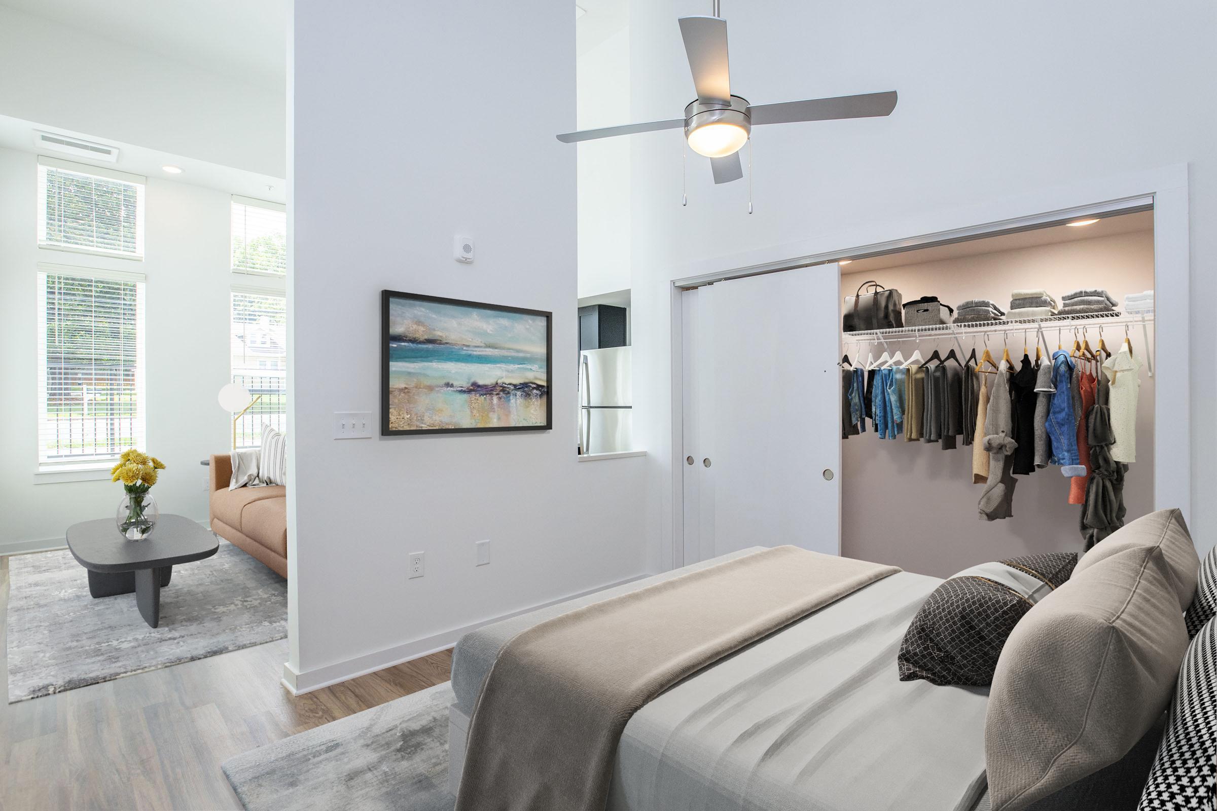 A stylish bedroom featuring a neatly made bed with decorative pillows, a ceiling fan, and a closet with organized clothing. In the background, large windows allow natural light, and a cozy living area with a sofa and modern decor can be seen. A painting hangs on the wall above the bed.