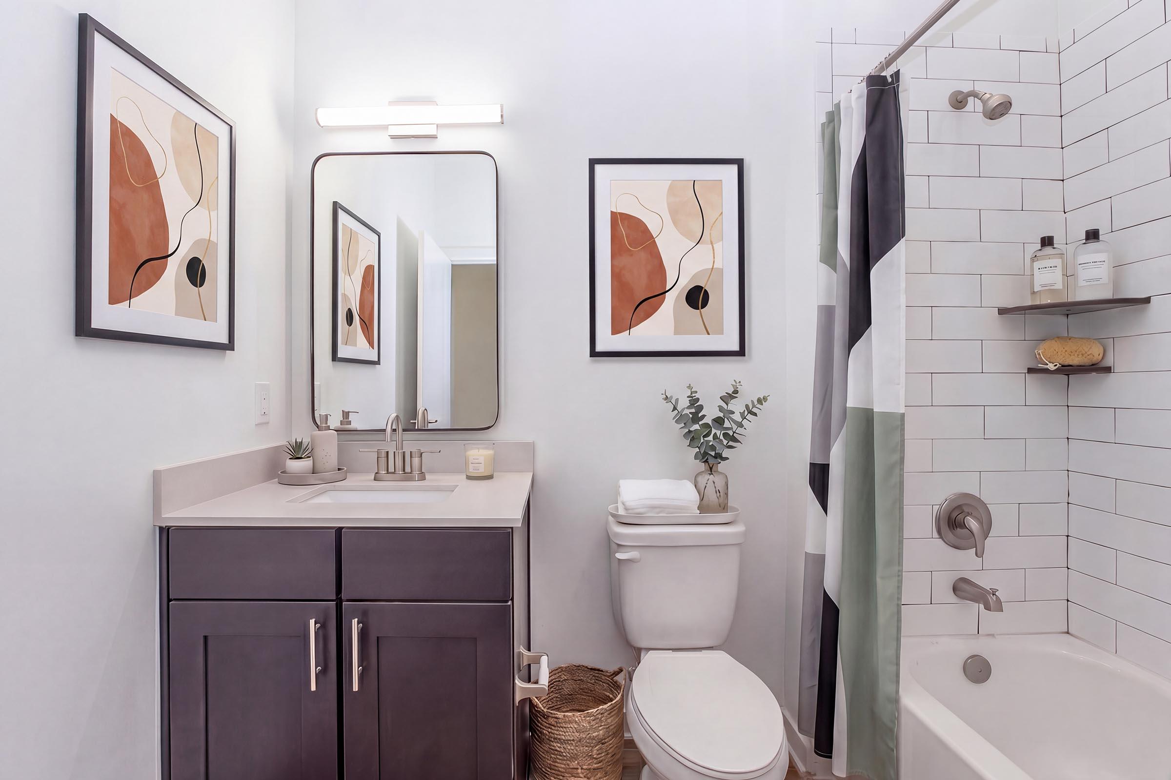 A modern bathroom featuring a sleek dark vanity with a countertop sink, a large mirror above, and framed abstract artwork on the walls. The space includes a white tiled shower with a black and green shower curtain, a toilet, and decorative elements like a small plant and a basket. Natural light brightens the room.