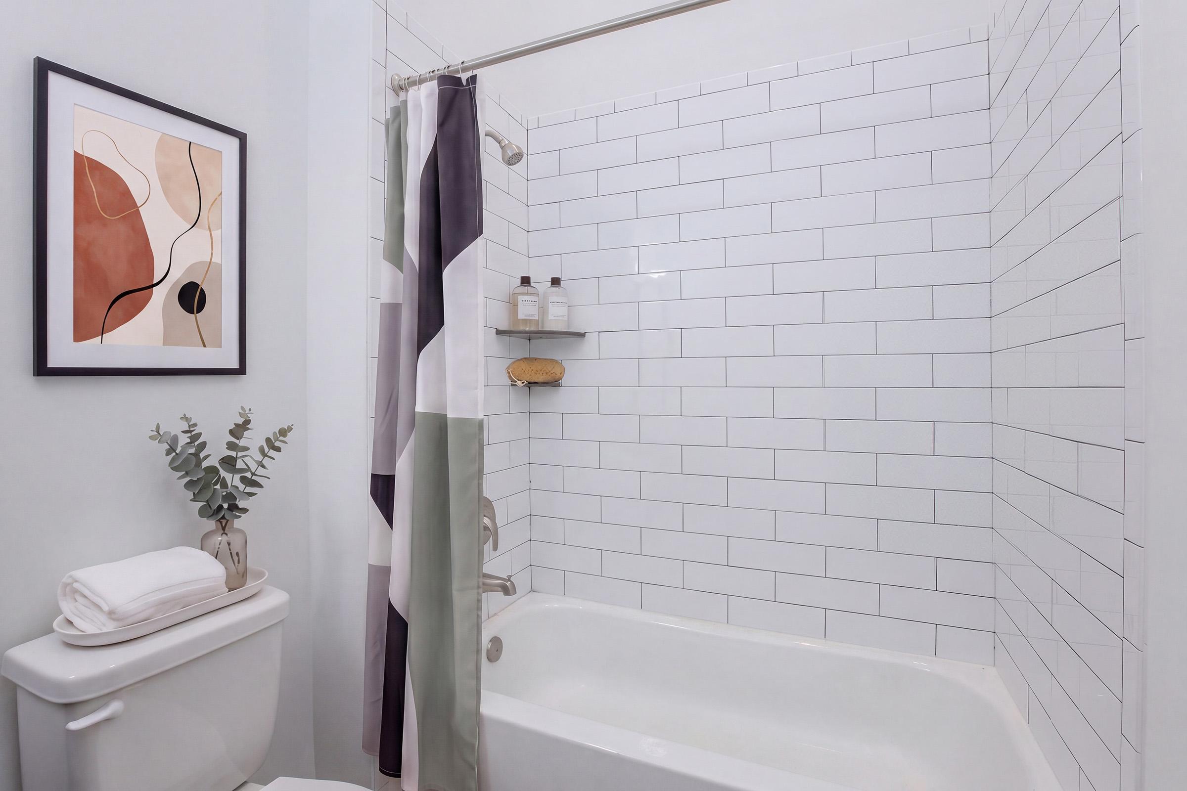 A modern bathroom featuring a clean, white tiled shower area with a glass door. There’s a white bathtub, a toilet, and a plant in a vase. The shower curtain has geometric patterns in muted colors. On the wall, there's an abstract art piece with warm tones. Shelves hold bath products, adding a touch of decor.