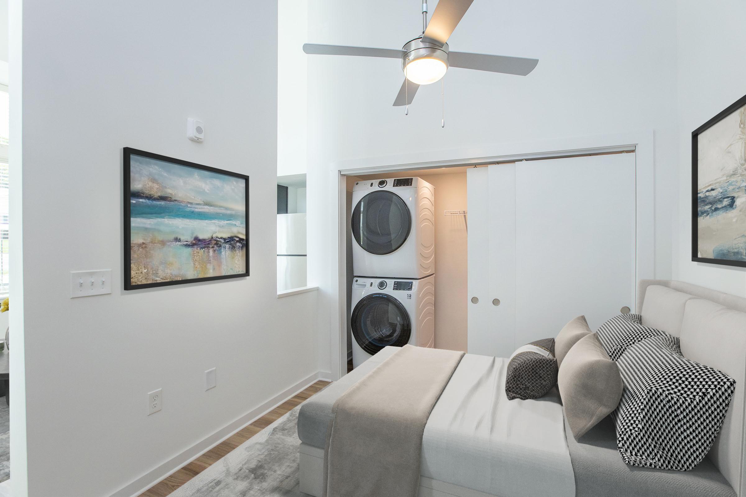 A modern bedroom with a light color palette, featuring a bed with decorative pillows, a ceiling fan, and a wall-mounted art piece. There is a washer and dryer stacked in a closet, and large windows allowing natural light to illuminate the space. The decor is minimal and contemporary.