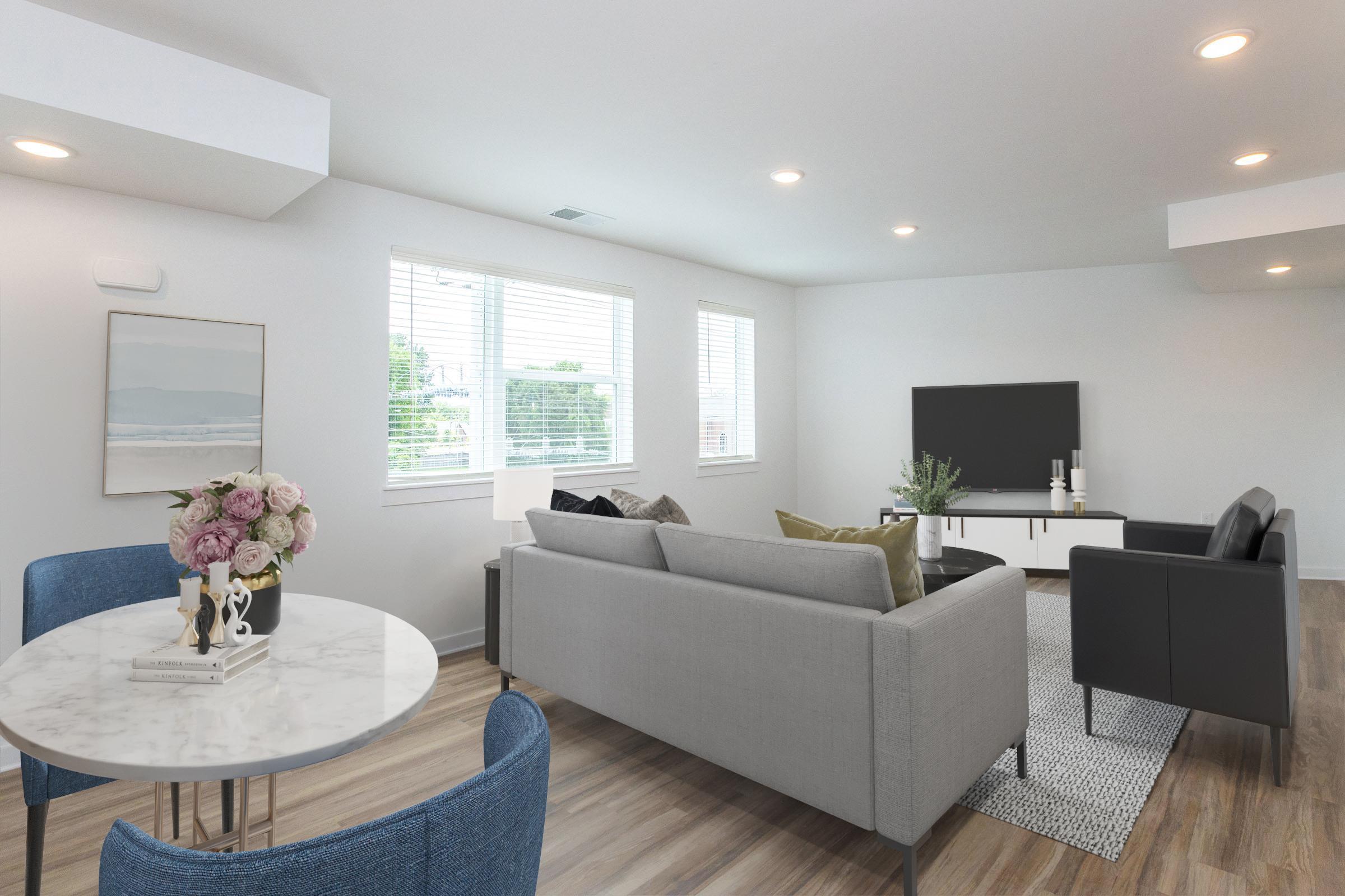 A modern, bright living room featuring a gray sofa, a black TV on a white entertainment unit, and a round marble dining table with blue chairs. There are large windows allowing natural light, and decorative elements like flowers on the table and a painting on the wall.