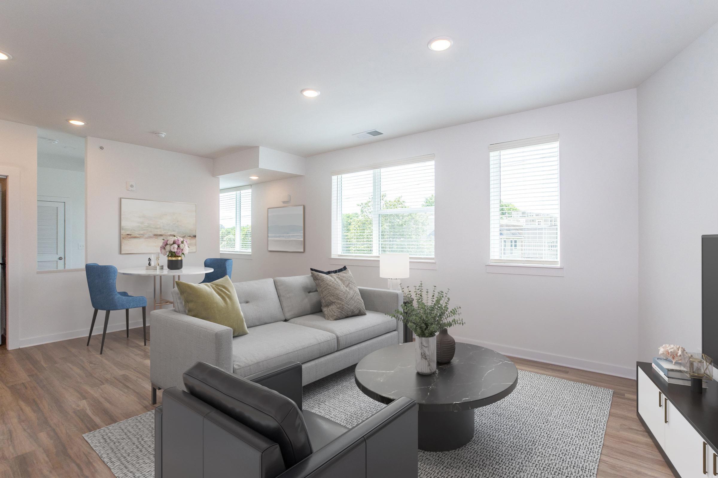 Bright and modern living room featuring a light gray sofa with decorative pillows, a round marble coffee table, and a black armchair. Large windows provide natural light, and a small dining table with chairs is visible in the background. Decorative plants and artwork add a cozy touch to the space.