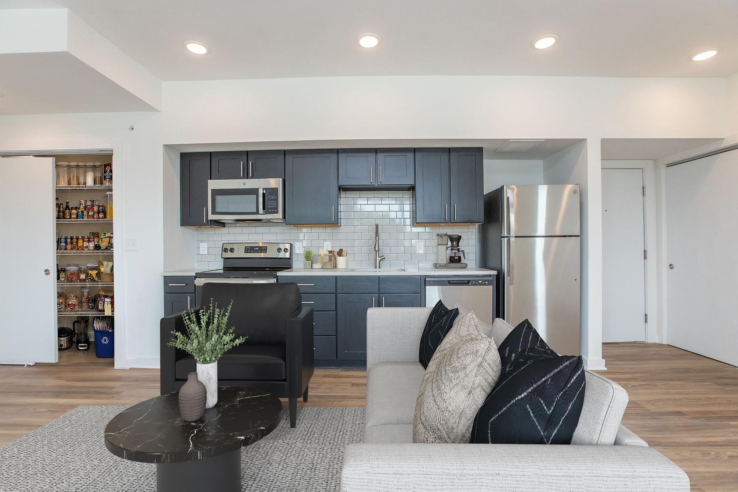 A modern kitchen and living area with navy blue cabinets, stainless steel appliances, and a pantry. The open layout includes a cozy gray sofa with decorative pillows and a black coffee table. Natural light fills the space, highlighting the wood-look flooring and neutral walls.