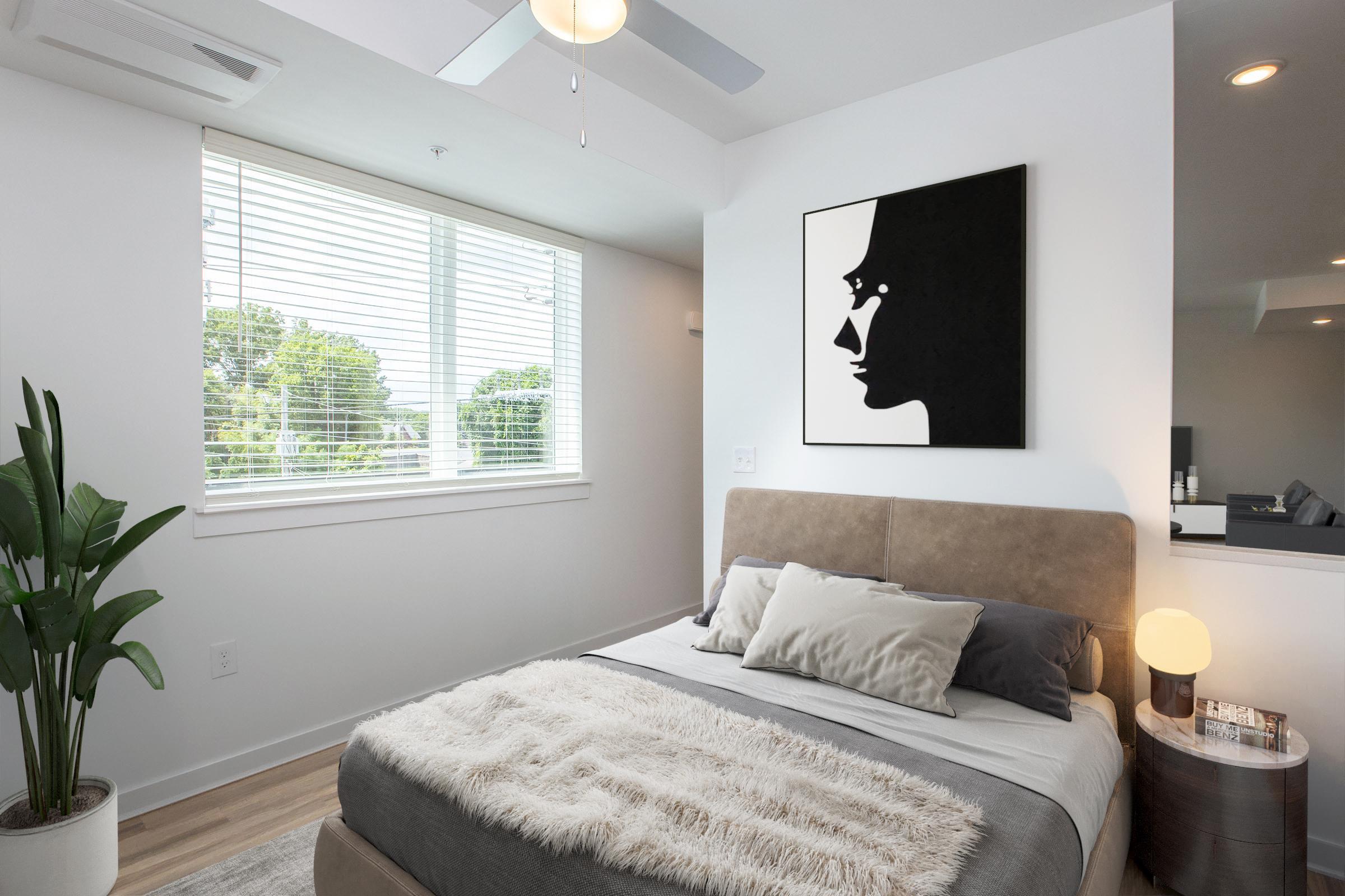 A modern bedroom featuring a queen-sized bed with grey and white bedding, a large wall art piece of a black and white profile silhouette, a small round bedside table with a lamp, and a large window with blinds allowing natural light to fill the room. A potted plant is placed beside the bed.