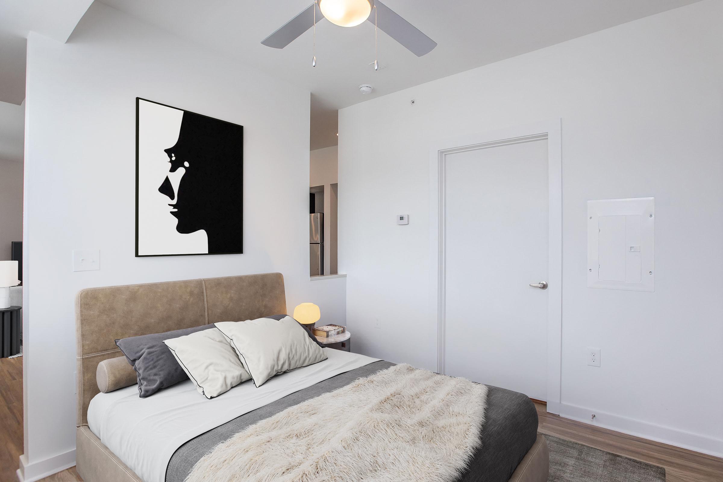 A modern bedroom featuring a beige bed with decorative pillows and a textured throw. A minimalist black and white artwork of a profile hangs on the wall above the bed. The room has a ceiling fan and a light fixture, with a door leading to another space. Neutral walls and wooden flooring complete the contemporary aesthetic.