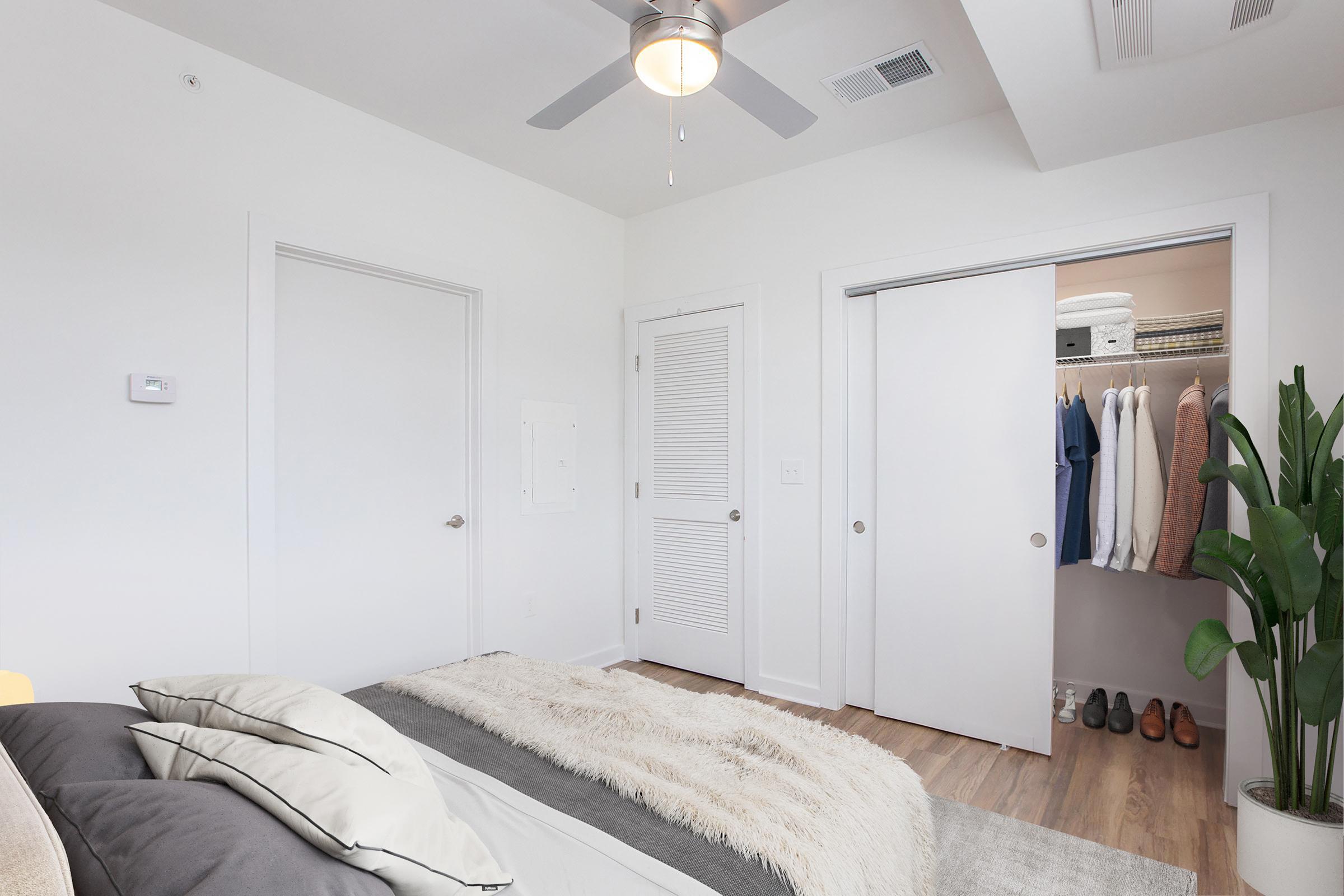 A cozy bedroom featuring a bed with decorative pillows and a plush throw blanket. There's a ceiling fan above, white walls, and a door leading to a closet with organized clothing. A green plant adds a touch of nature to the room. Natural light enhances the inviting atmosphere.