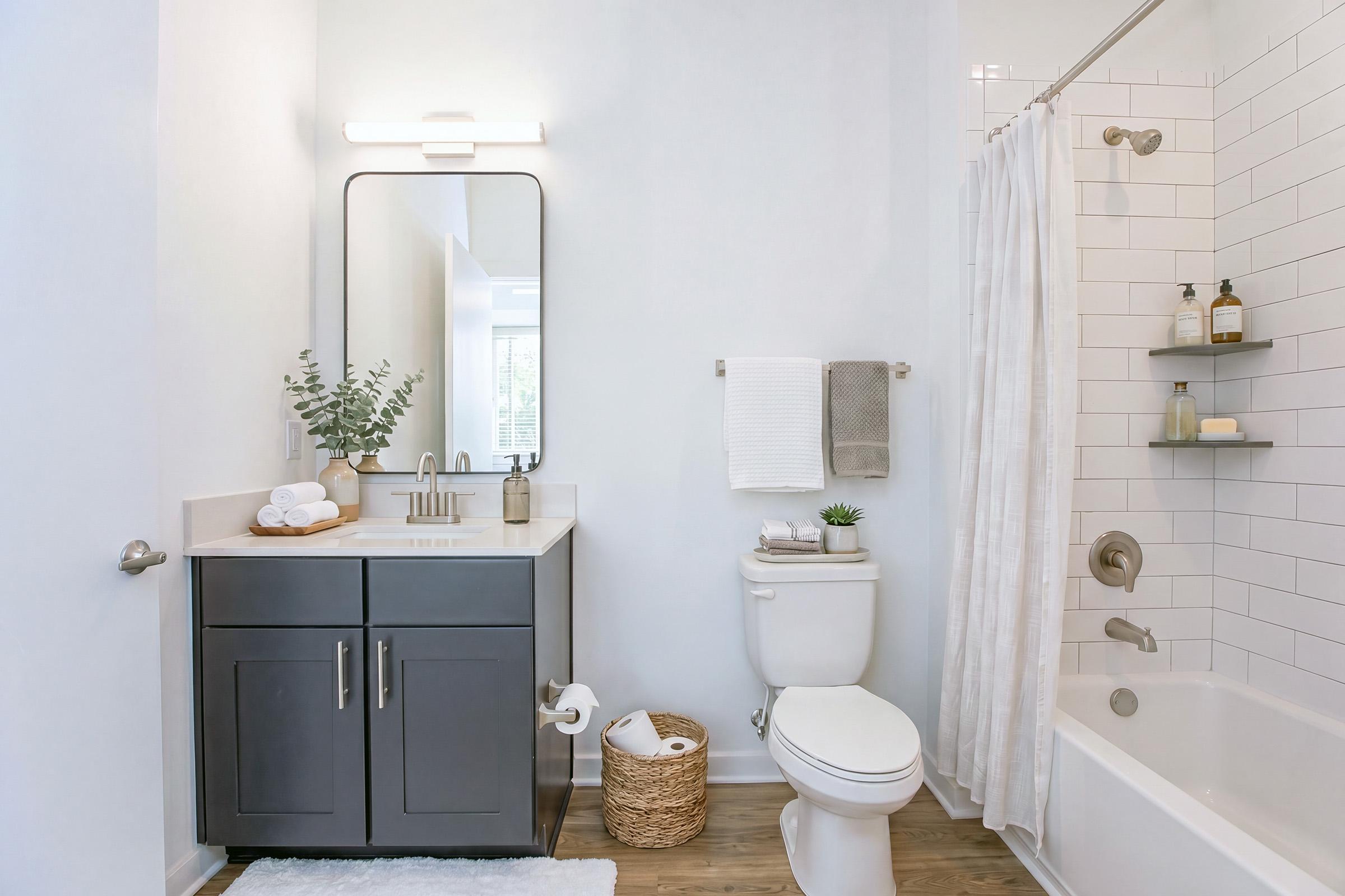 A modern bathroom featuring a white tiled shower with a clear curtain, a dark vanity with a sink and a large mirror, a toilet, and shelving with toiletries. Decorative elements include a small indoor plant, rolled towels, and a woven basket on the floor. The light-colored walls enhance the bright, airy atmosphere.