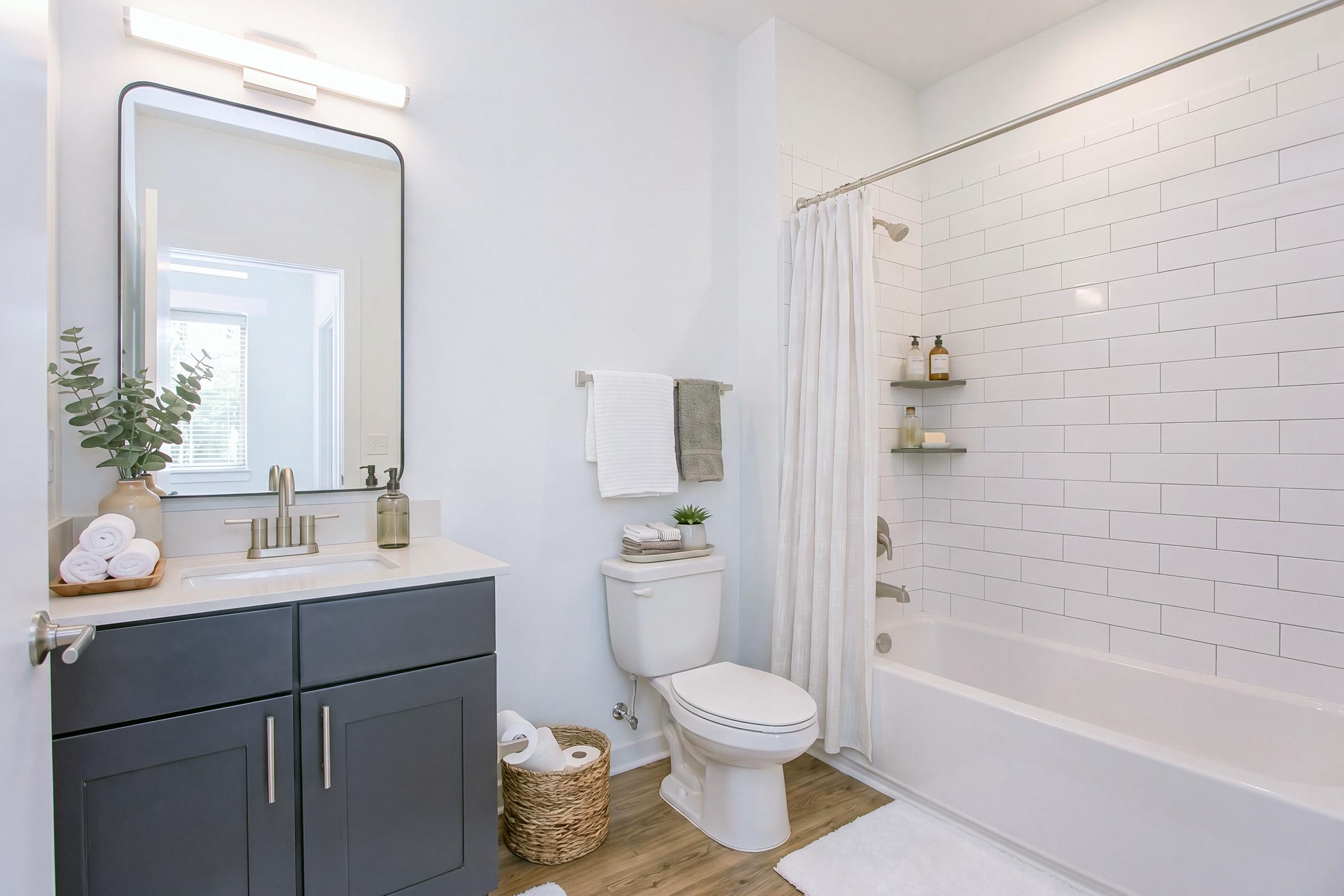 A modern bathroom featuring a white tile shower with a bathtub, a gray vanity with a large mirror, and a toilet. Decorative elements include a plant, neatly stacked towels, and a woven basket. The lighting is bright, creating a clean and inviting atmosphere.