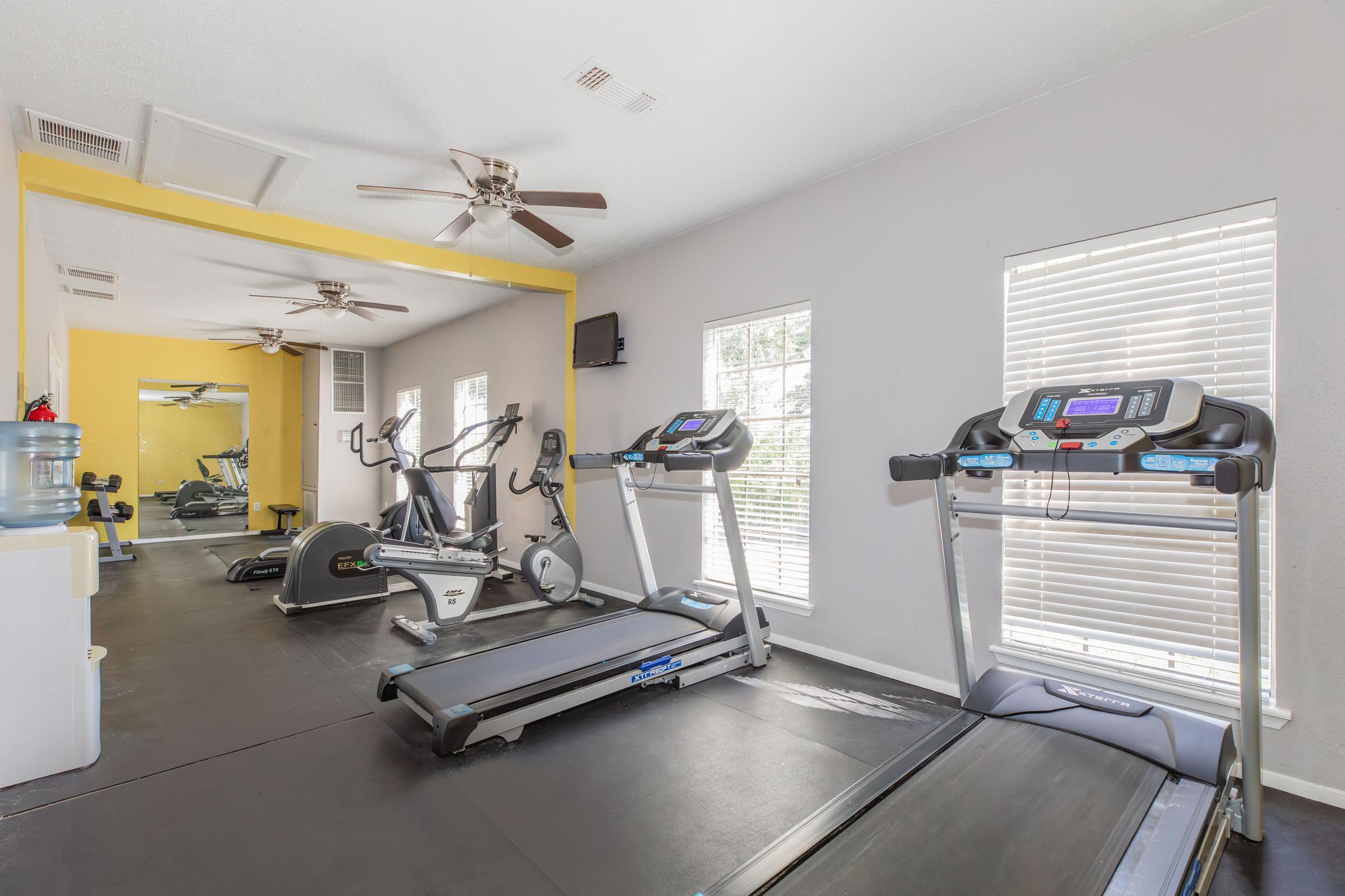 A bright, well-equipped gym featuring two treadmills and an elliptical machine. The space has large windows for natural light, a water cooler, and ceiling fans. The walls are painted in light gray with a yellow accent, creating an inviting workout atmosphere.