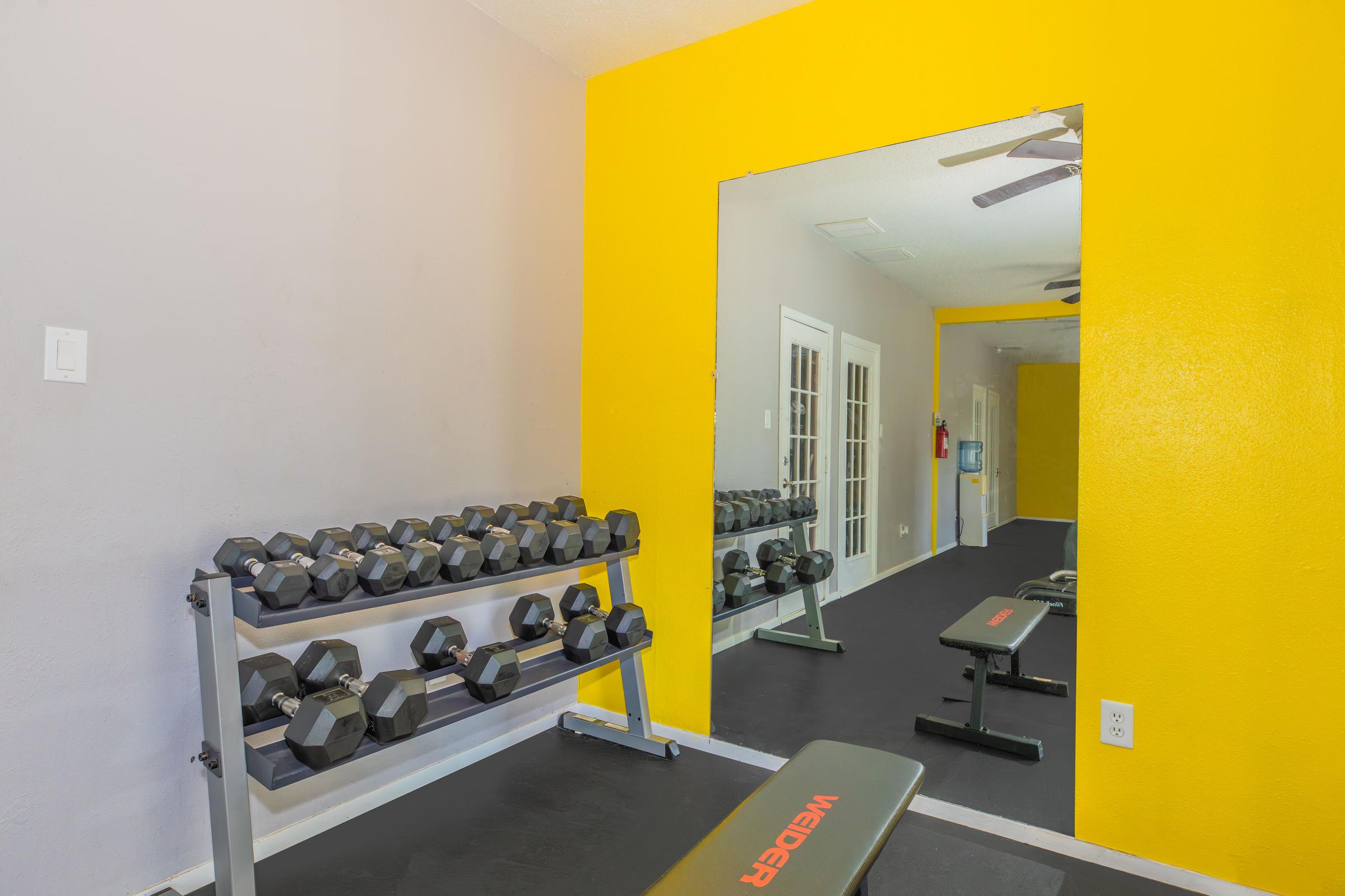 A well-lit home gym featuring a rack of dumbbells, a workout bench, and a large mirror on a bright yellow wall. The flooring is dark, and there are windows providing natural light. The space is functional and tidy, with a few workout accessories visible.