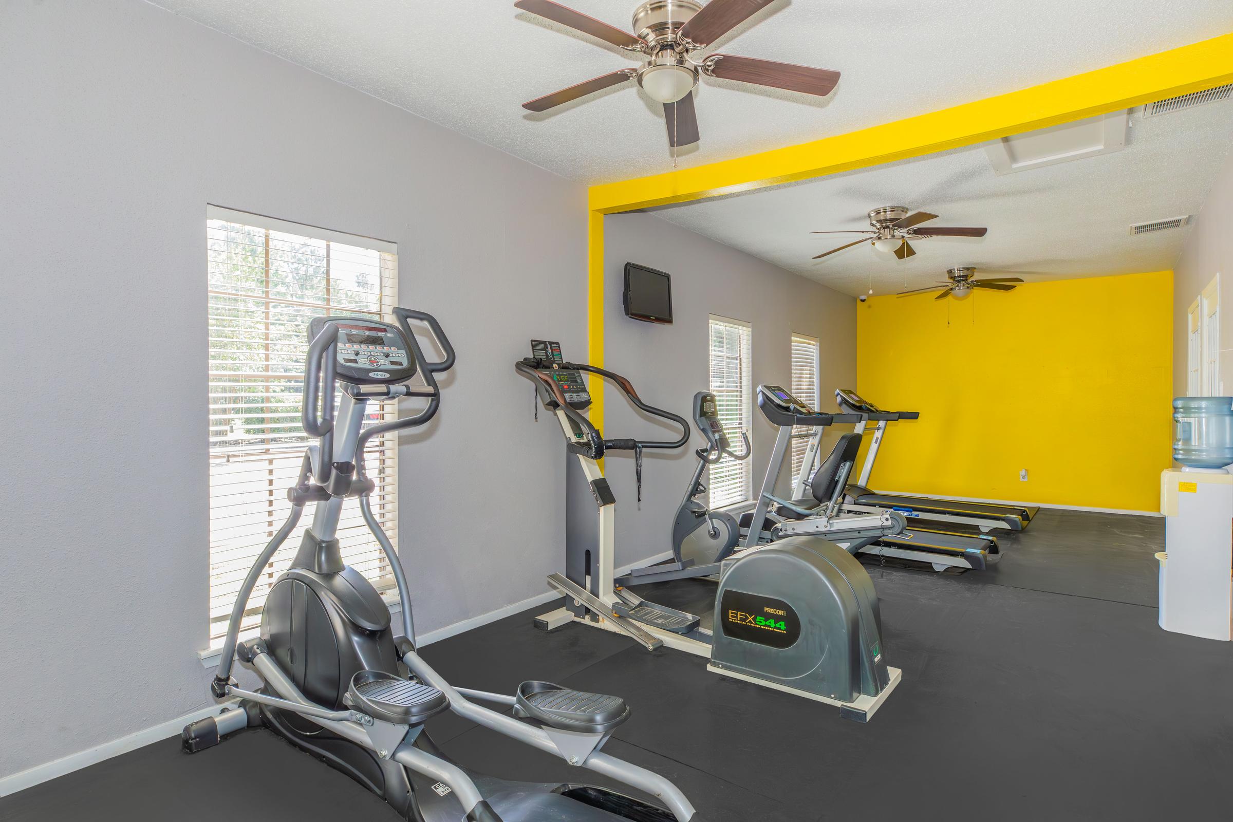 A bright and spacious gym area featuring exercise equipment, including elliptical machines and a rowing machine. The walls are painted in light gray and a vibrant yellow accent wall. Ceiling fans are installed, and there is a water cooler visible. Large windows allow natural light to fill the room.