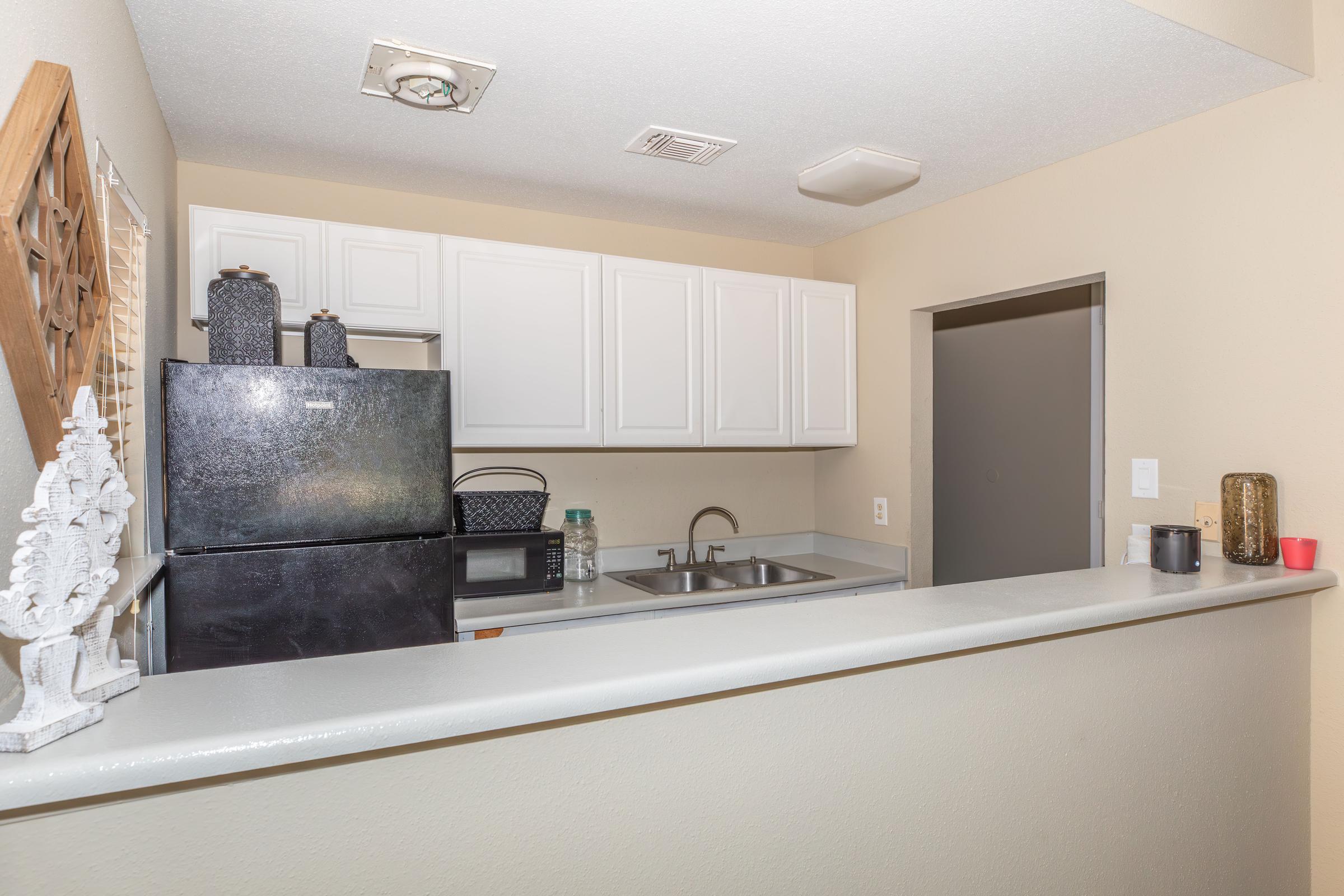 A view of a modern kitchen featuring white cabinetry, a black refrigerator, a sink with a faucet, and a microwave. The kitchen is open to a living area with a light beige wall finish. Decorative items are placed on the counter, adding subtle touches to the space.