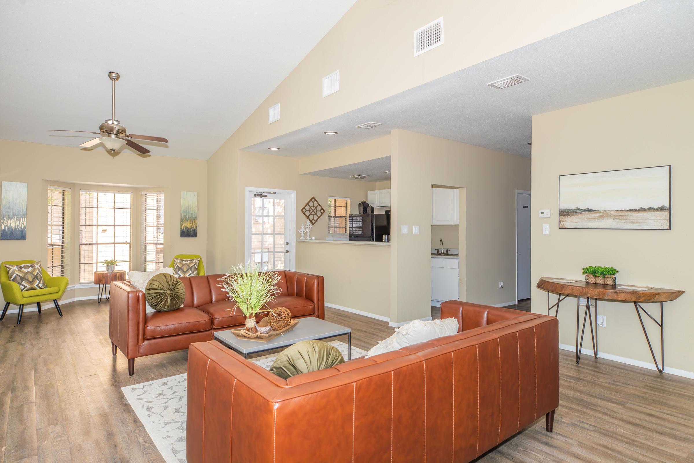 A spacious living room featuring two brown leather sofas, a coffee table, and a decorative plant. Large windows provide natural light, and there are cozy chairs near the window. The room has a modern aesthetic with beige walls and wooden flooring, and an open kitchen can be seen in the background.
