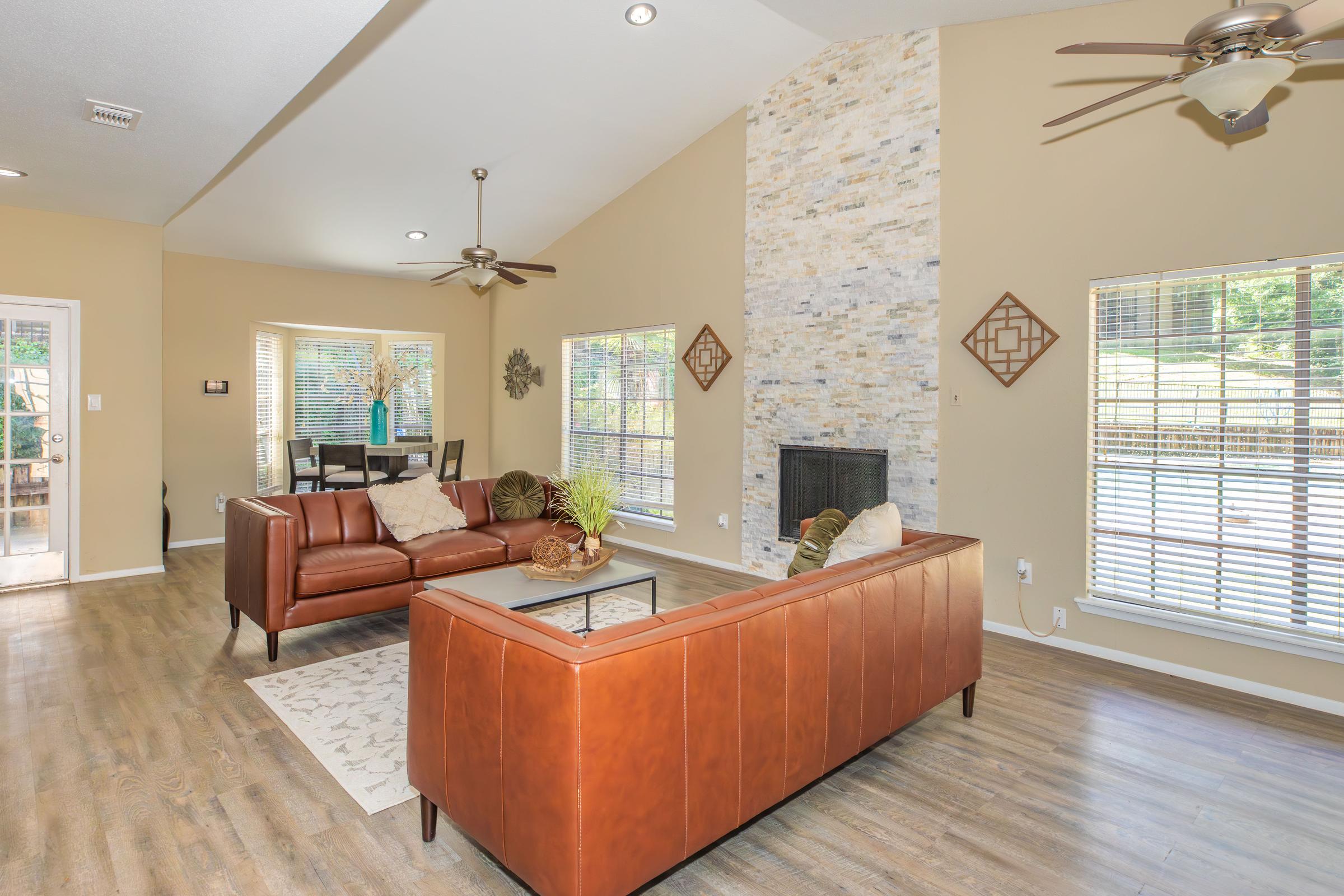 A spacious living room featuring a tan-colored sofa, a coffee table, and a stone accent wall. Large windows allow natural light, and a dining area with a brown table is visible in the background. The flooring is a warm wood tone, and there are decorative items like plants and wall art.