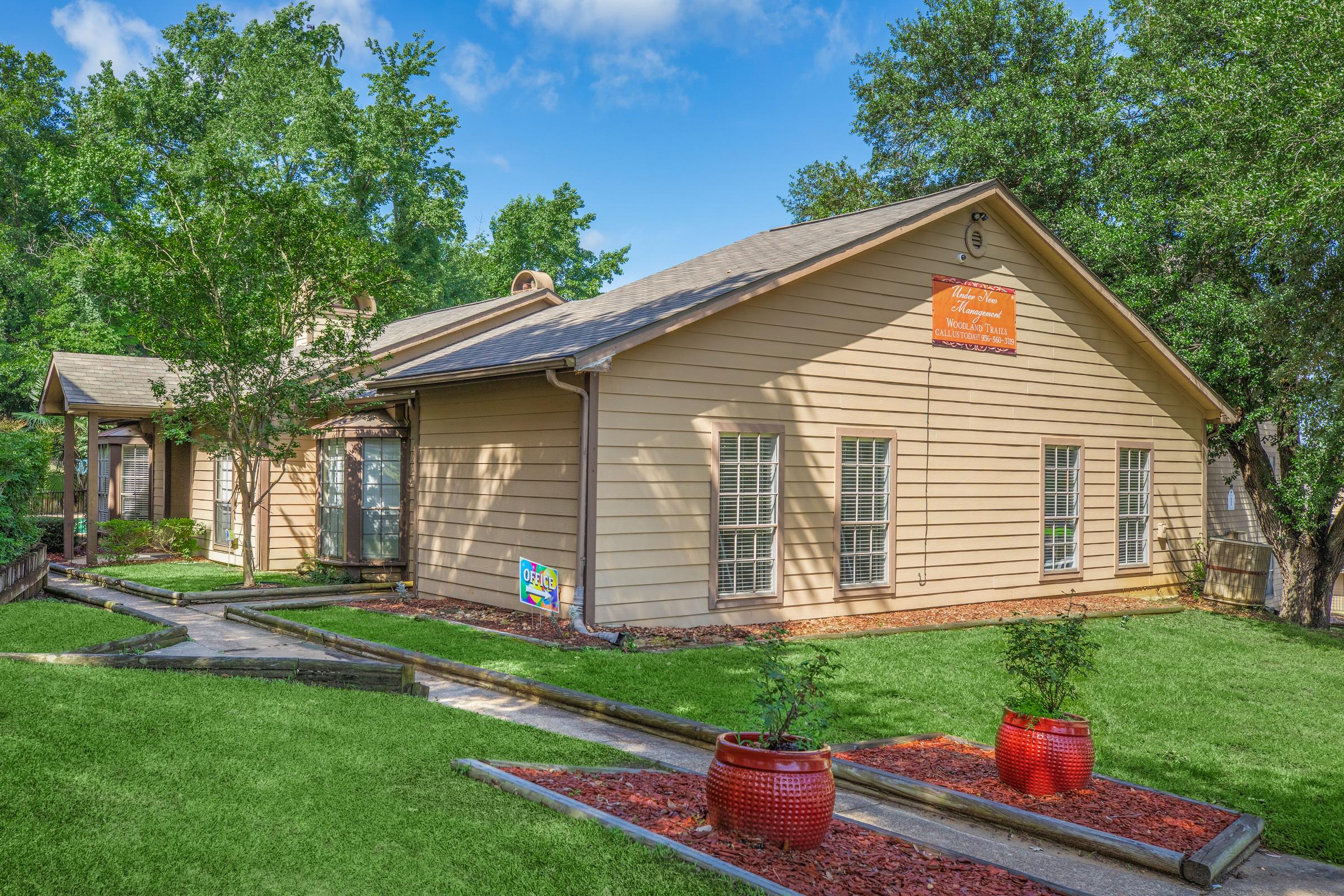 A single-story beige house with a sloped roof and a wooden sign hanging over the entrance. The front yard features green grass, red flower pots, and a pathway leading to the door. Surrounding trees provide shade, and the sky is a bright blue with a few clouds.