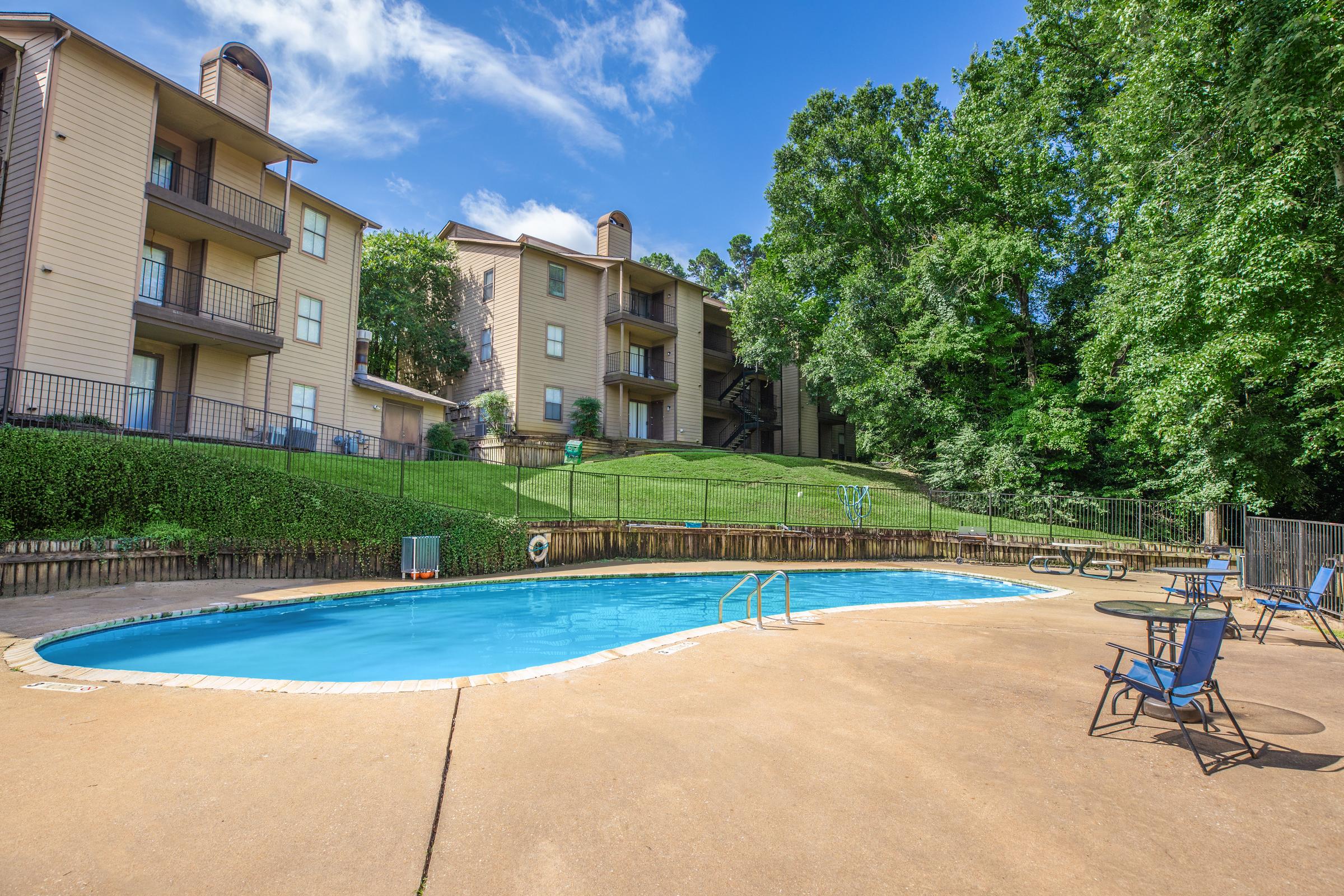 A tranquil outdoor swimming pool surrounded by lounge chairs, with well-maintained grass and trees in the background. The pool area is adjacent to an apartment building with multiple levels, under a clear blue sky with a few clouds.