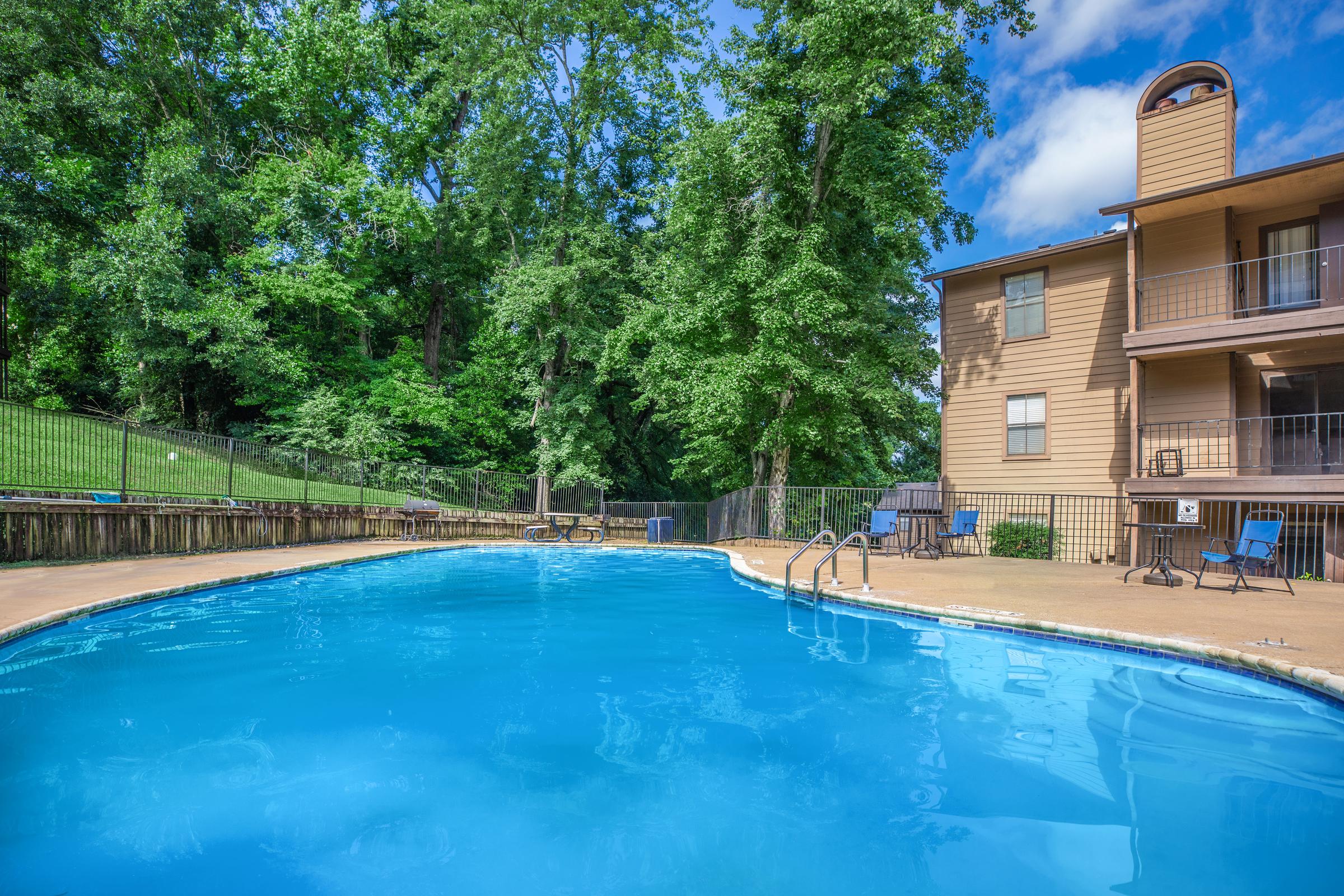 A bright and inviting swimming pool surrounded by lush green trees, with a residential building nearby. Sunlight reflects off the clear blue water, creating a serene atmosphere. Poolside seating is available, and a well-maintained fence encloses the area, enhancing the outdoor experience.