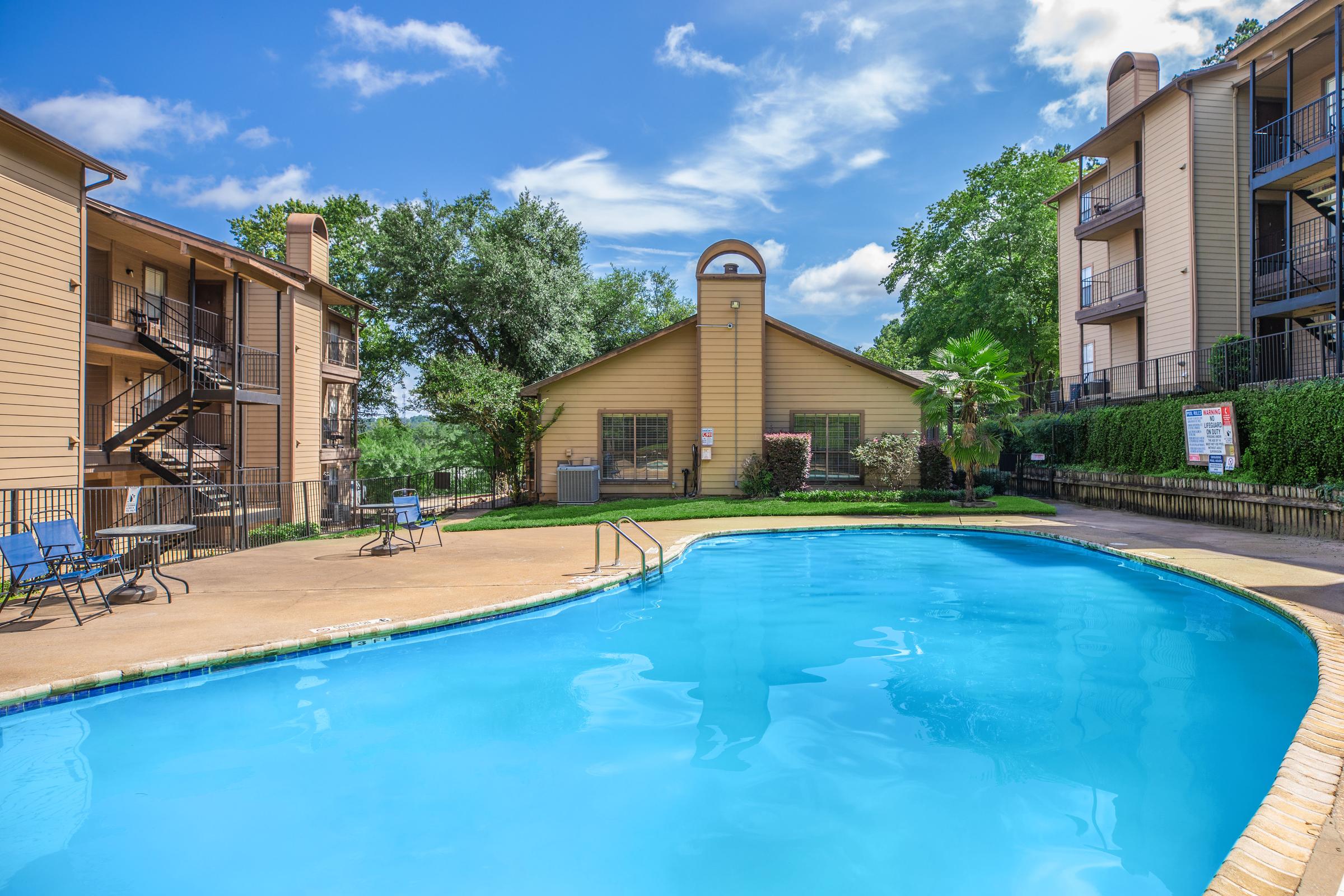 A sunny outdoor swimming pool surrounded by landscaped greenery and buildings. The pool features a clear blue water surface, with lounge chairs nearby. In the background, there is a clubhouse or community center, along with well-maintained landscaping and a blue sky with scattered clouds.