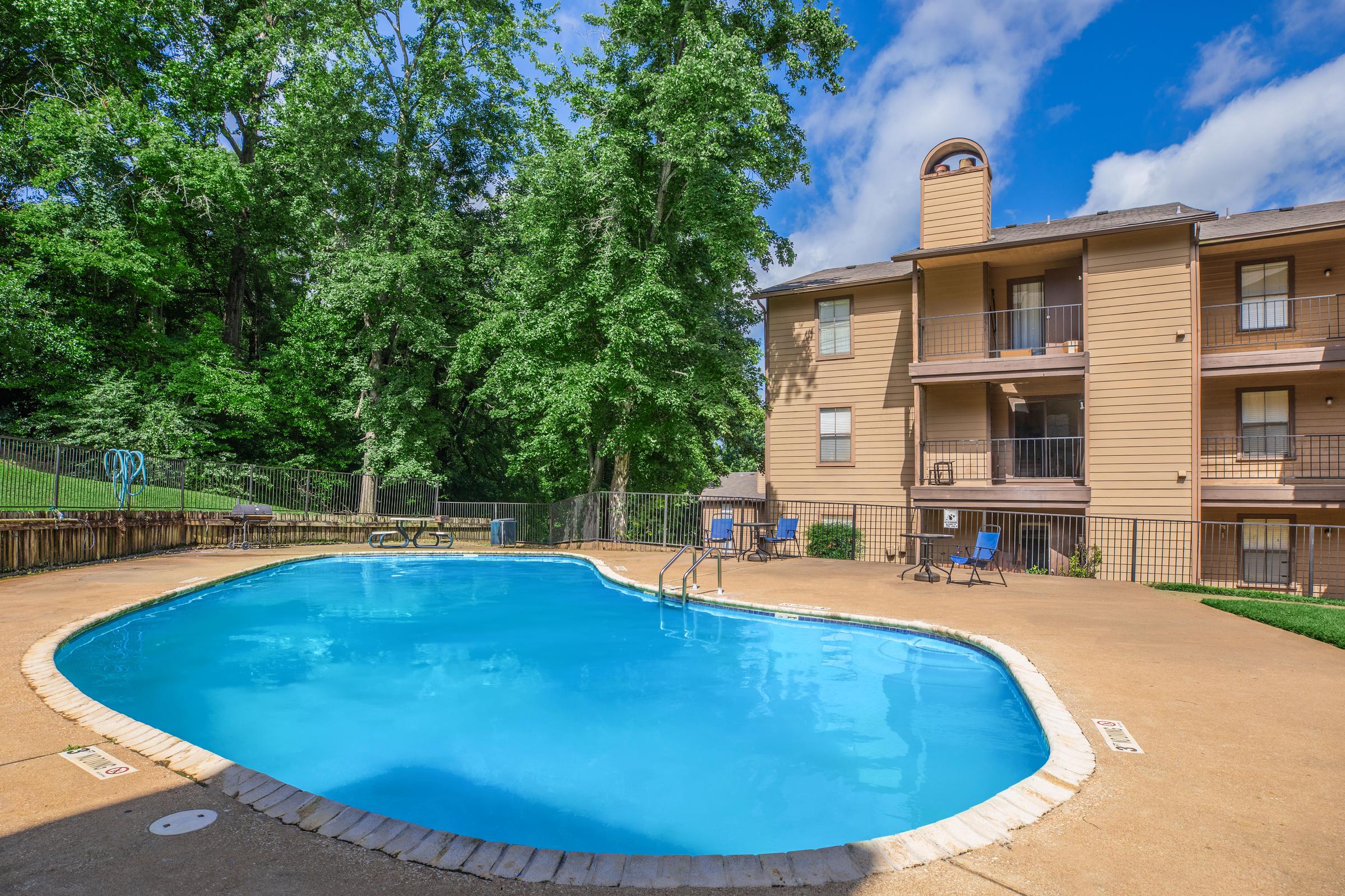 A bright blue swimming pool surrounded by a sunny outdoor area, with lounge chairs nearby. In the background, a three-story apartment building is framed by lush green trees under a clear blue sky, creating a relaxed and inviting atmosphere.