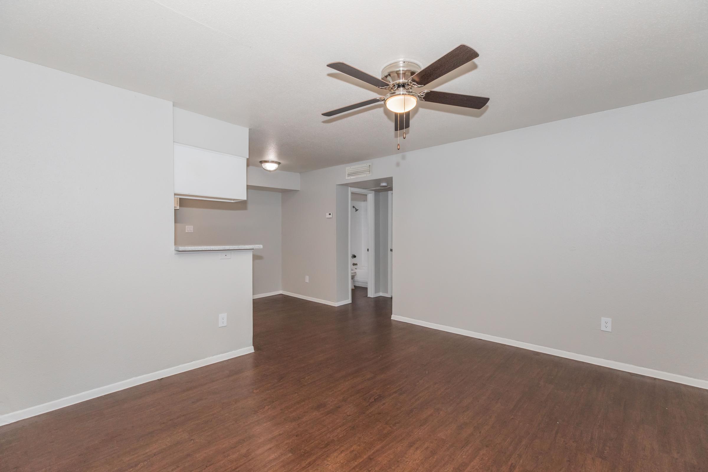 A spacious, well-lit living area featuring a ceiling fan, light-colored walls, and a wood-style floor. A partial view of a kitchen area is visible in the background, along with a doorway leading to a bathroom. The overall ambiance is modern and inviting.