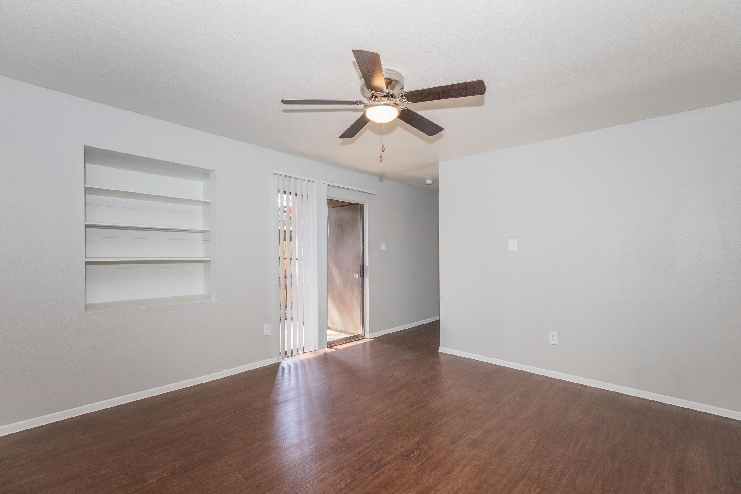 Spacious interior of an apartment featuring a ceiling fan, light-colored walls, and laminate flooring. There are built-in shelves on one side and a door leading outside, providing natural light to the room. The layout is open and uncluttered, creating a welcoming atmosphere.