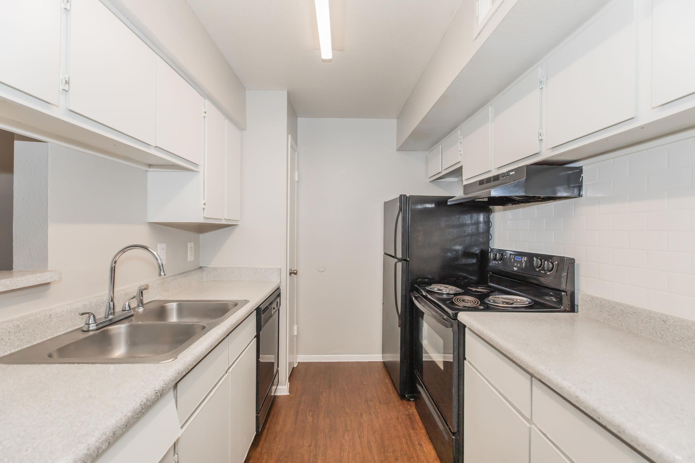 Modern kitchen featuring light-colored cabinetry, a dual sink, granite-style countertops, and a black refrigerator and oven. The space has a clean aesthetic with white tile backsplash and wooden flooring, providing a functional and inviting cooking area.