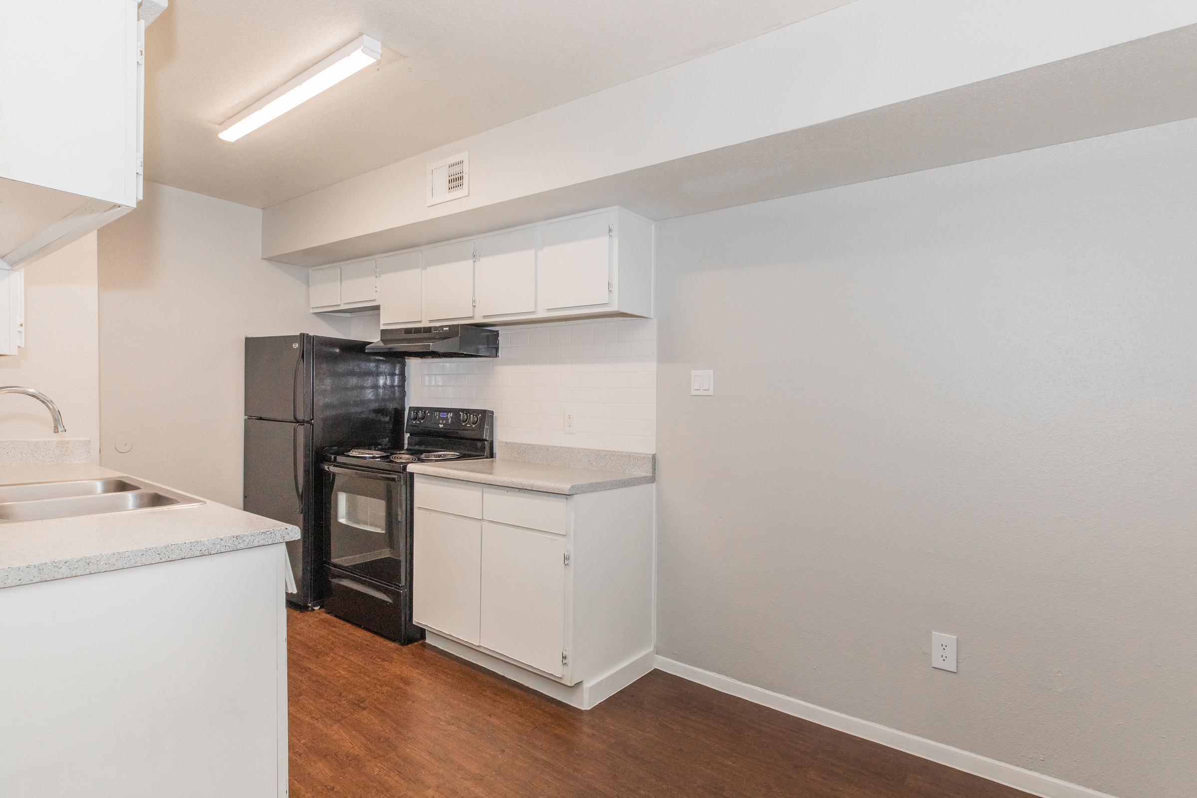 Modern kitchen with white cabinets, a black refrigerator, and a black stove. Countertops are a light gray, and the floor has a warm wood-like finish. The walls are painted a light gray, and there is overhead lighting. A sink is visible in the foreground.