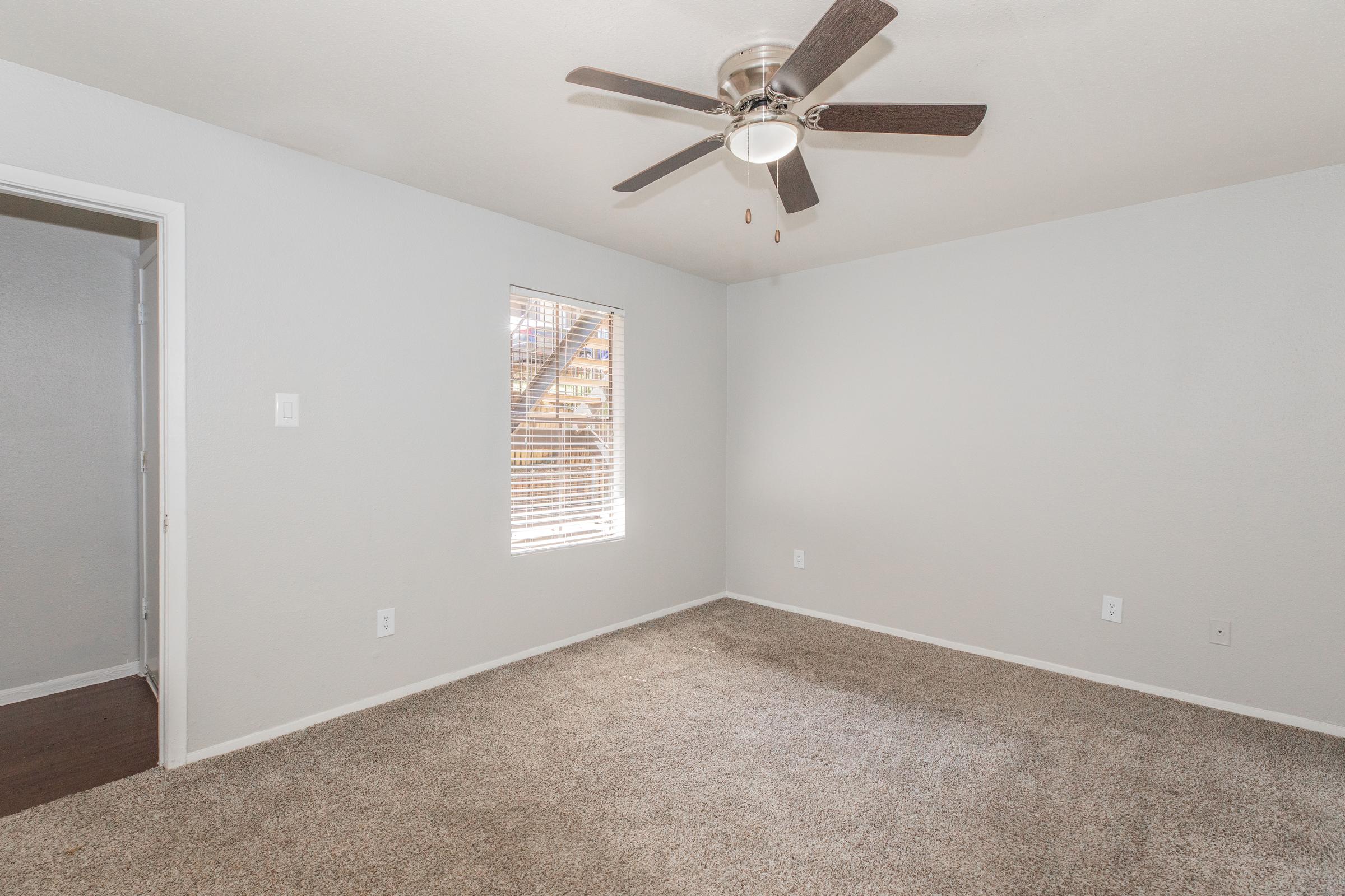 A spacious, empty room with light gray walls and beige carpet. A ceiling fan with wooden blades hangs in the center. There is a window with horizontal blinds allowing natural light to enter. A doorway leading to another room is visible on the left. The overall feel is clean and well-lit.