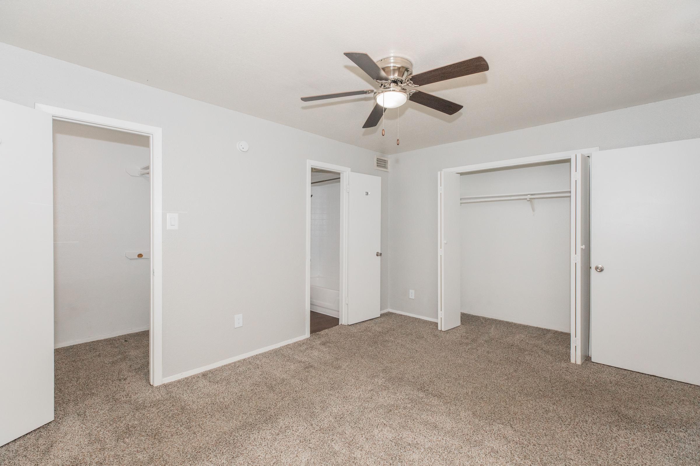 A spacious bedroom featuring light-colored walls, beige carpet, a ceiling fan, and two open doors leading to closets. The room is well-lit, with ample space for furniture and decor. A bathroom door is also visible in the background, enhancing the room's functionality.