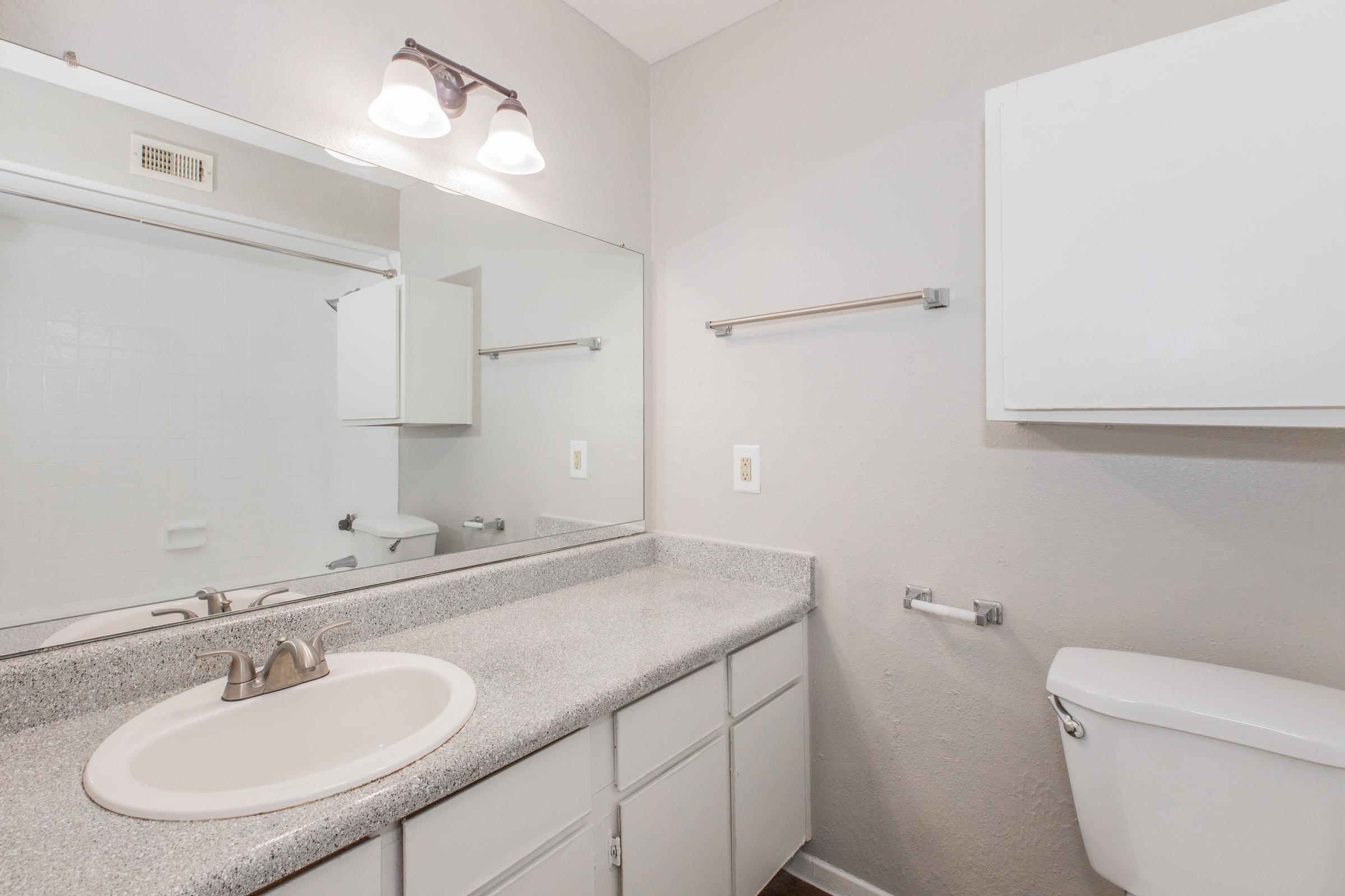 A clean and modern bathroom featuring a light gray countertop with a sink, a large mirror above it, white cabinetry, a towel bar, and a toilet. The walls are painted light gray, and there is a simple light fixture above the mirror. The overall design is minimalist and fresh.