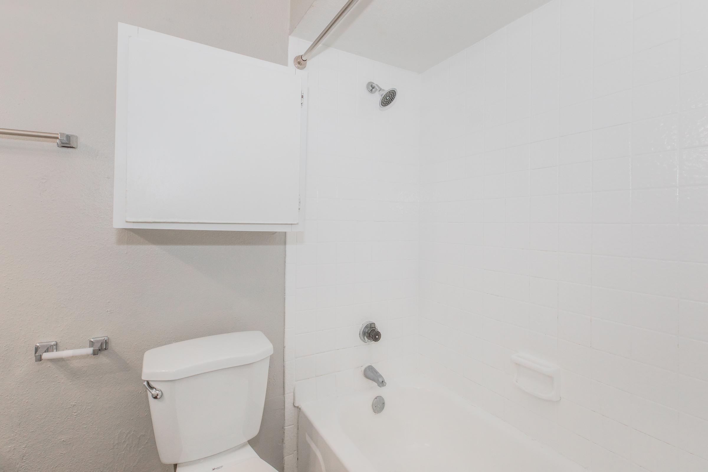 A clean bathroom scene featuring a white bathtub and a toilet. The walls are painted light gray, and there is a white showerhead and faucet. A small white cabinet is visible on the wall, providing storage. The overall appearance is simple and functional.