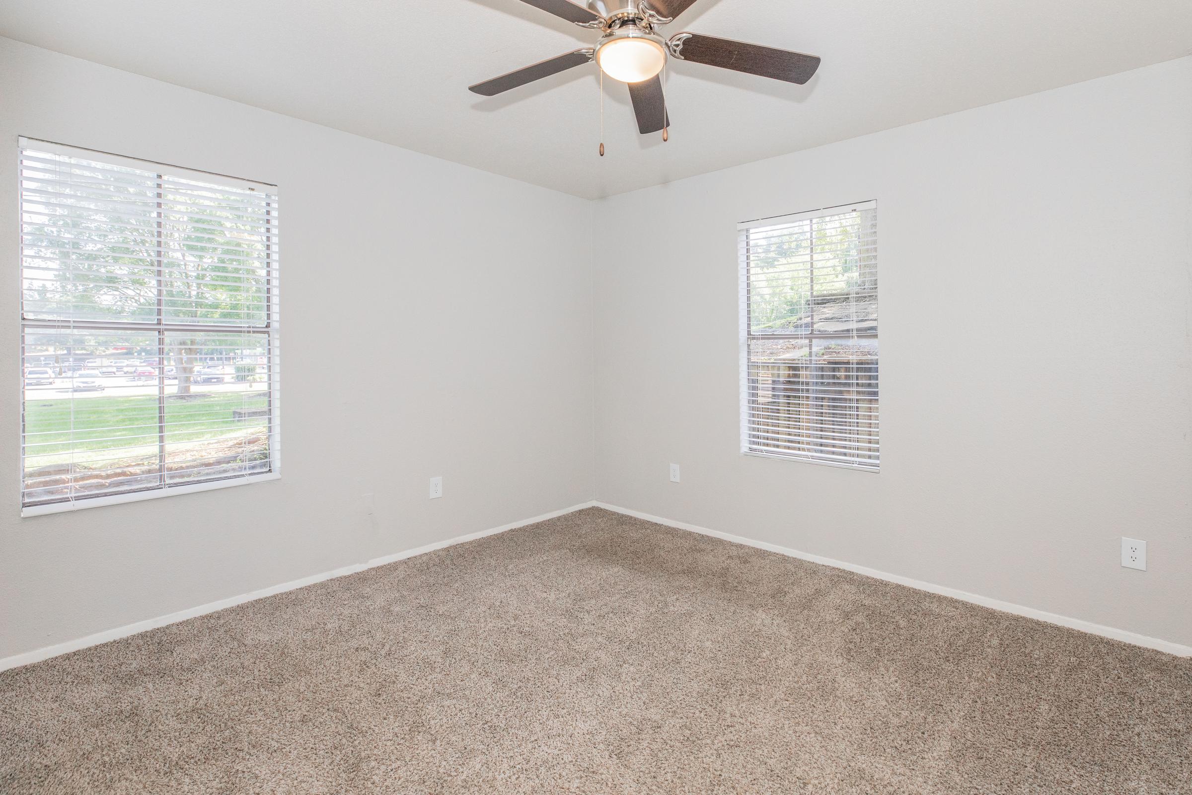A vacant room with light gray walls and beige carpet. It features two windows with blinds, allowing natural light to enter. A ceiling fan is mounted in the center of the room, creating an airy feel. The space is clean and ready for furniture arrangement.