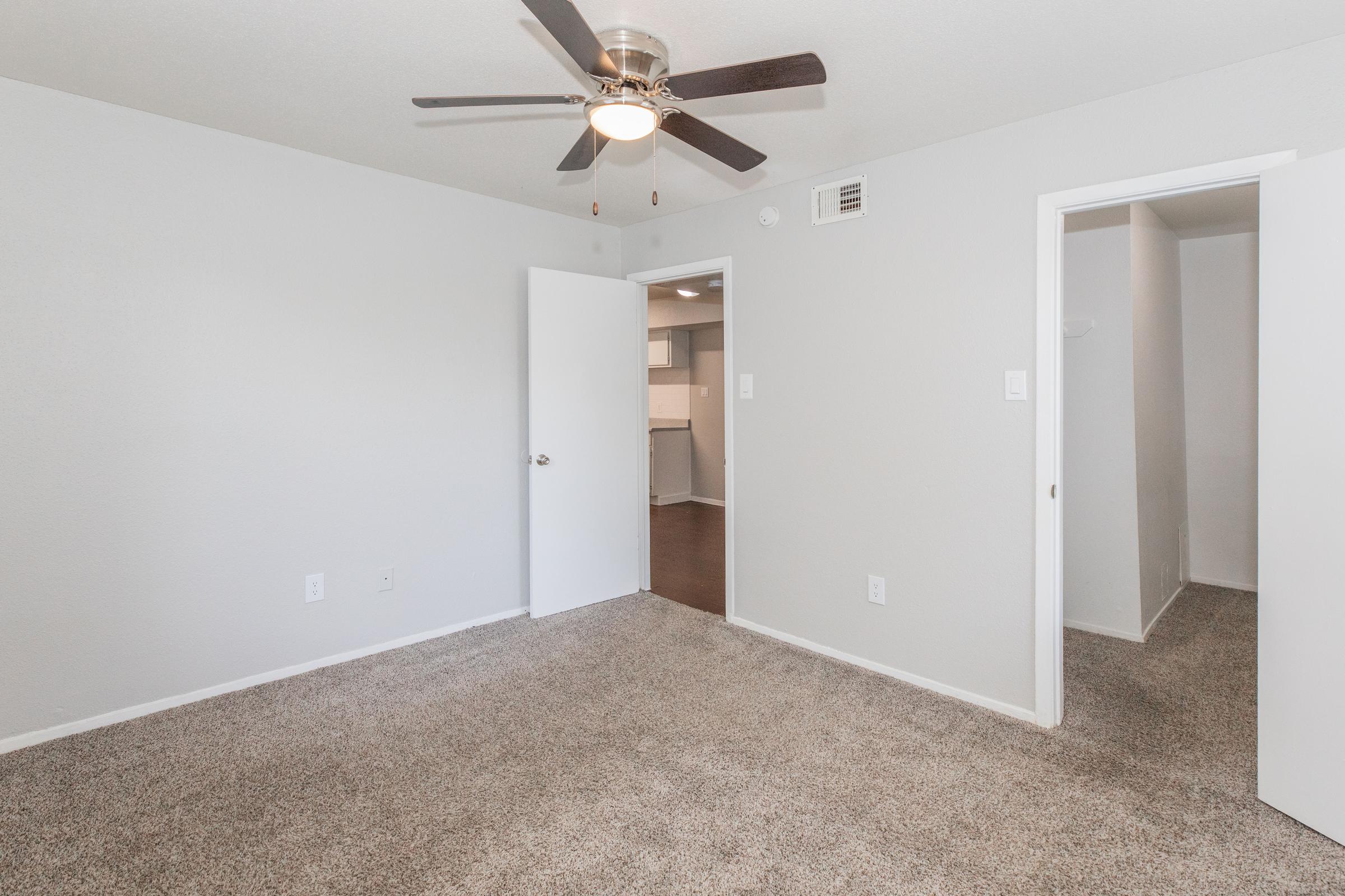 A spacious, well-lit room with beige carpet, featuring a ceiling fan. Two white doors are open, leading to adjacent areas. The walls are painted light gray, providing a neutral backdrop. The room appears clean and inviting, ideal for various uses such as a bedroom or office space.