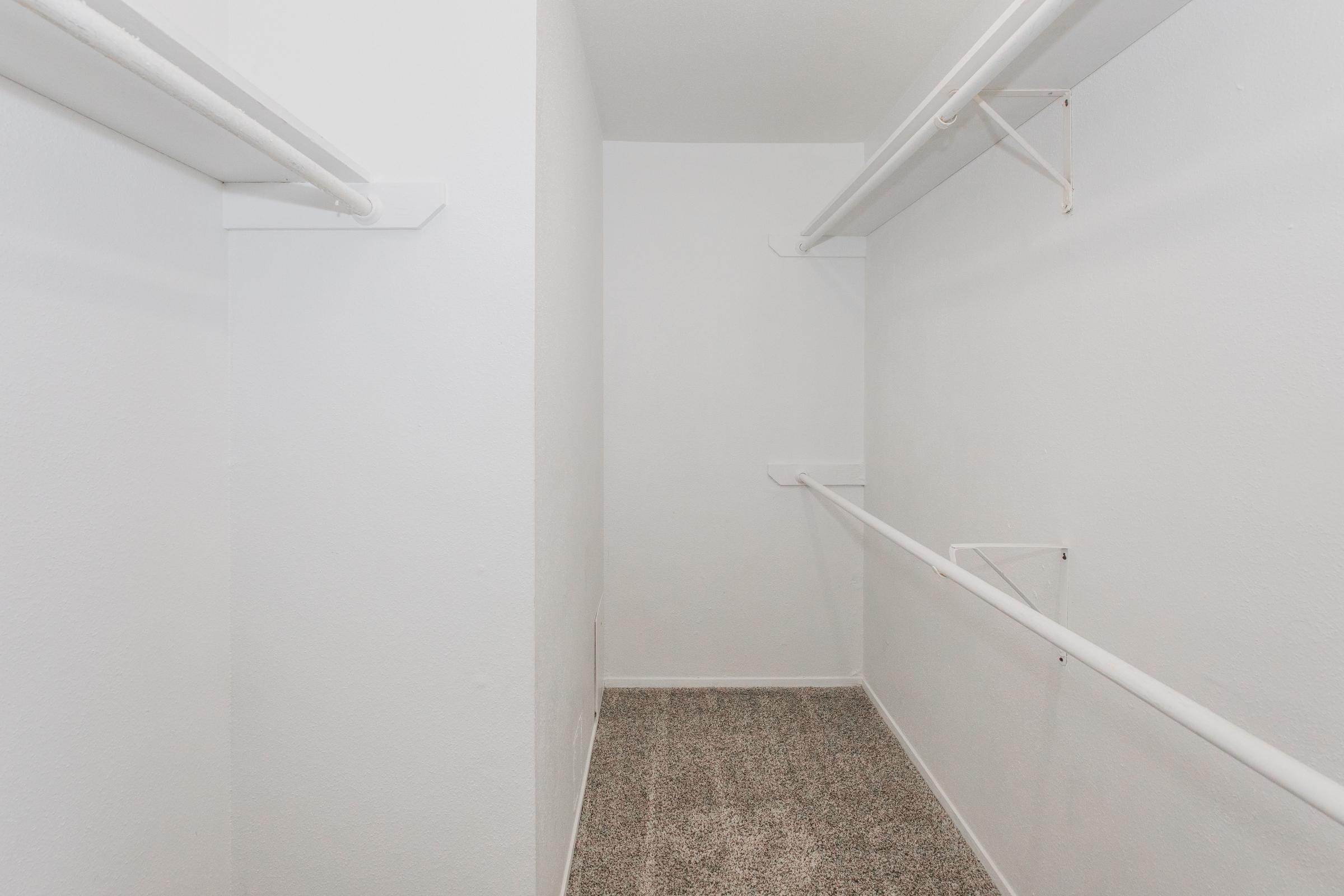 A spacious, empty closet featuring white walls and ceiling, with light-colored carpet flooring. There are horizontal rods for hanging clothes on both sides, and the overall appearance is clean and minimalist.